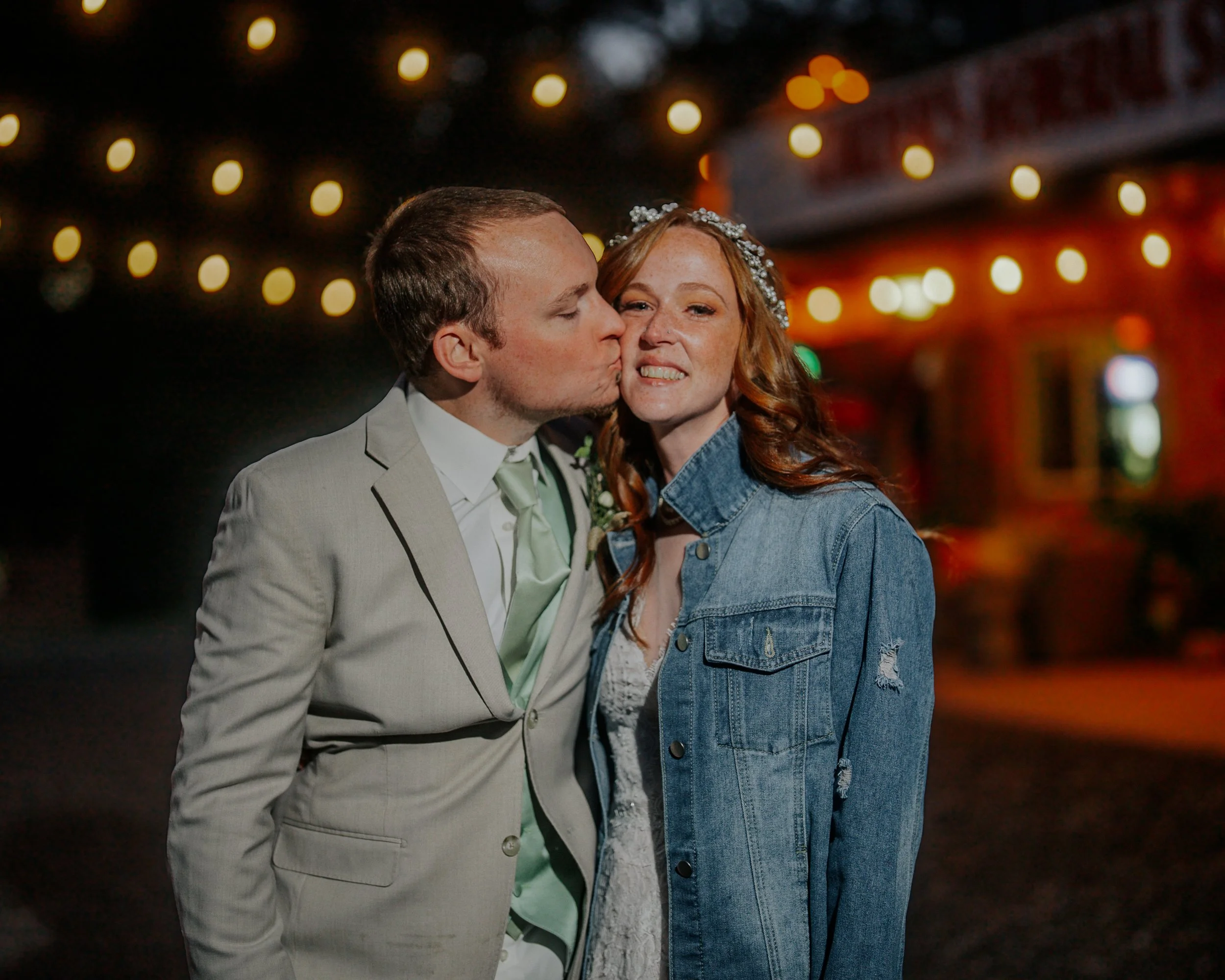 A man in a light-colored suit kisses a woman with red hair and a denim jacket on the cheek at night, with string lights in the background.
