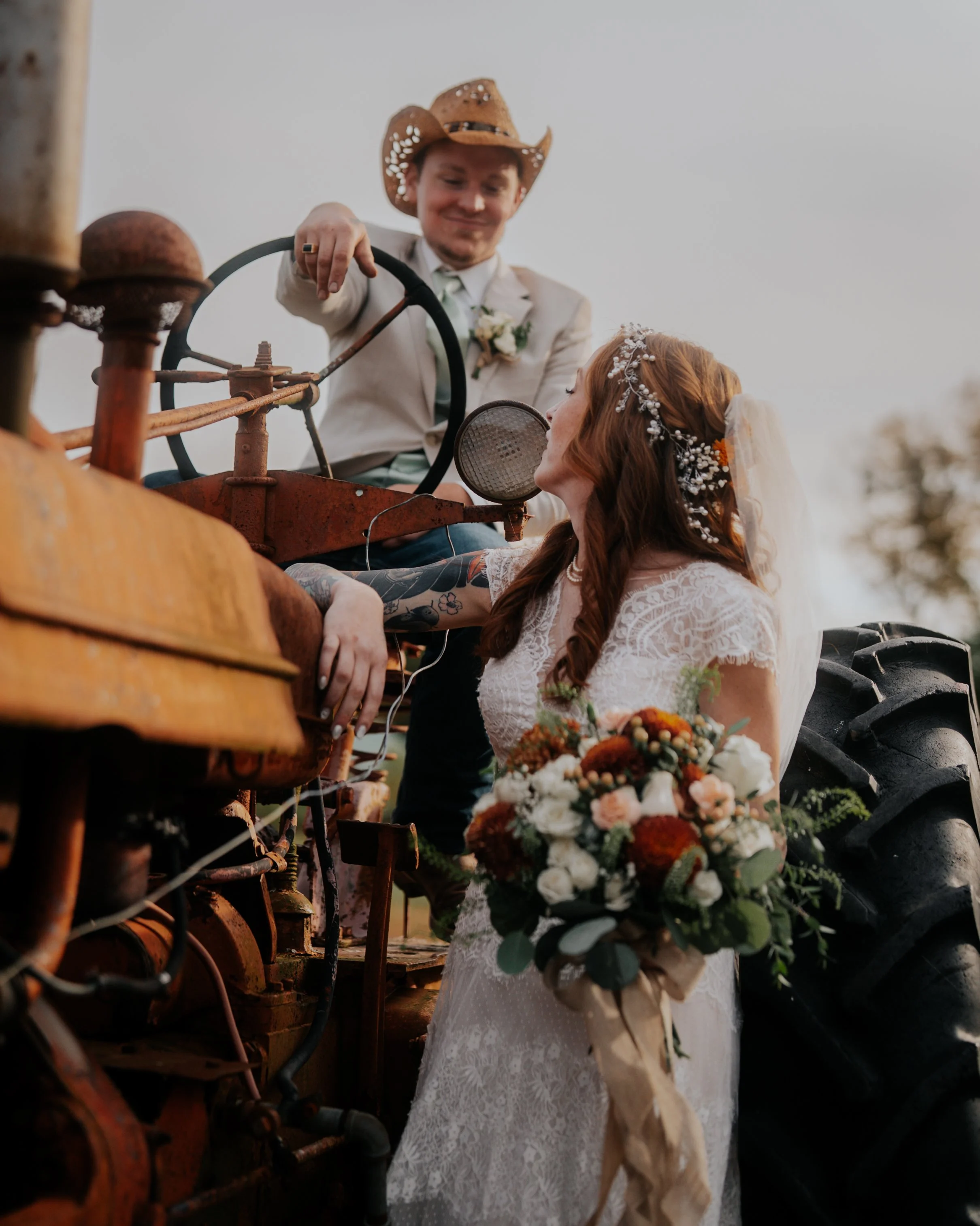 A bride in a white lace dress holding a bouquet of flowers looking up at a groom on an old tractor, both wearing cowboy hats, during a wedding photo shoot.
