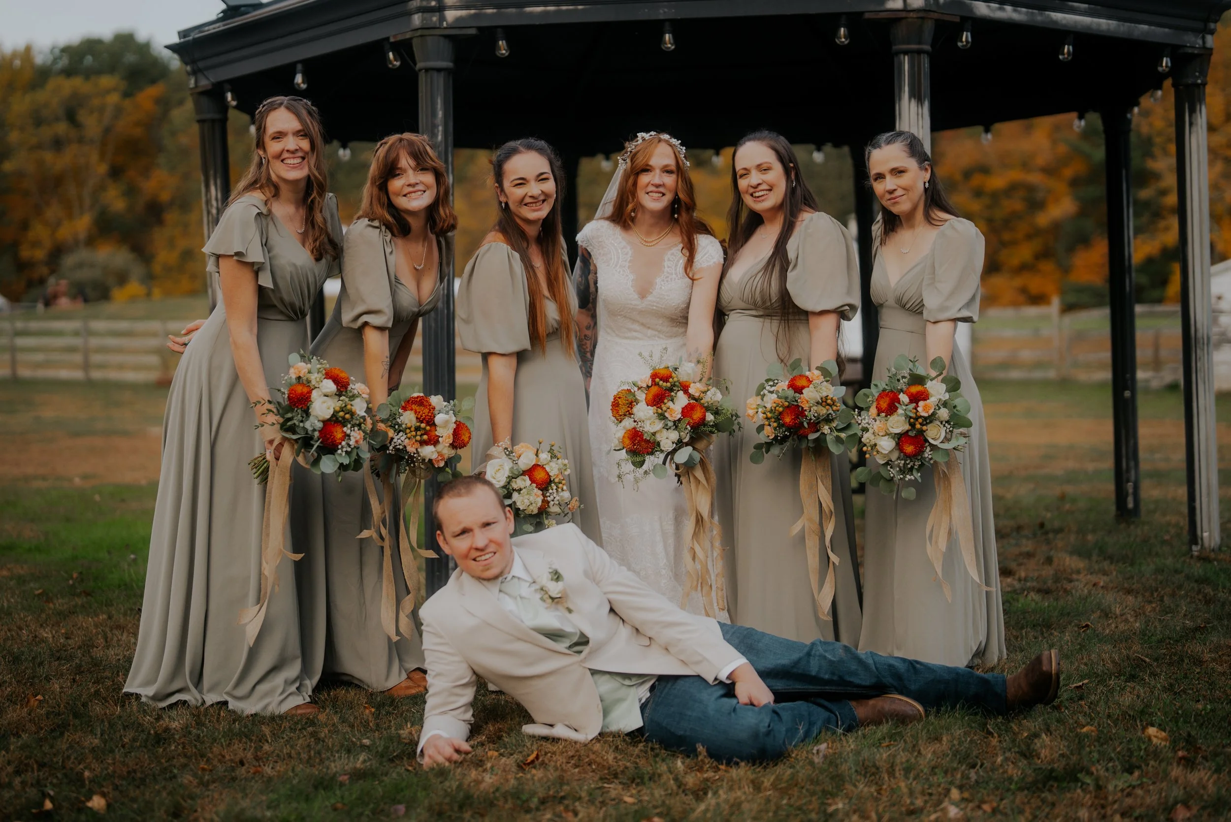 A group of seven women in matching light gray bridesmaid dresses and one bride in a white wedding dress standing under a gazebo outdoors, holding bouquets of flowers. A young man in a white jacket and dark jeans lies on the grass in front of them, sm