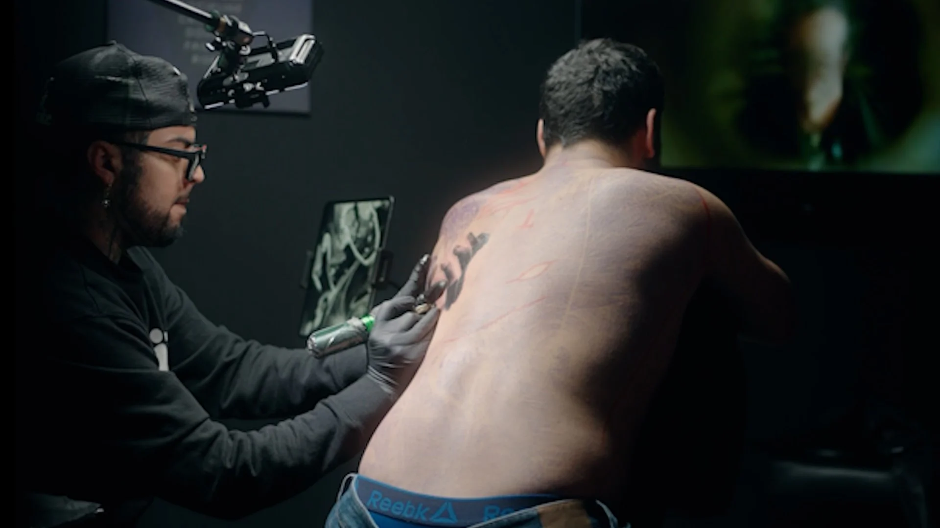 A tattoo artist in a black cap and glasses tattooing a man's back, who is shirtless and wearing Reebok shorts, in a dark studio environment.