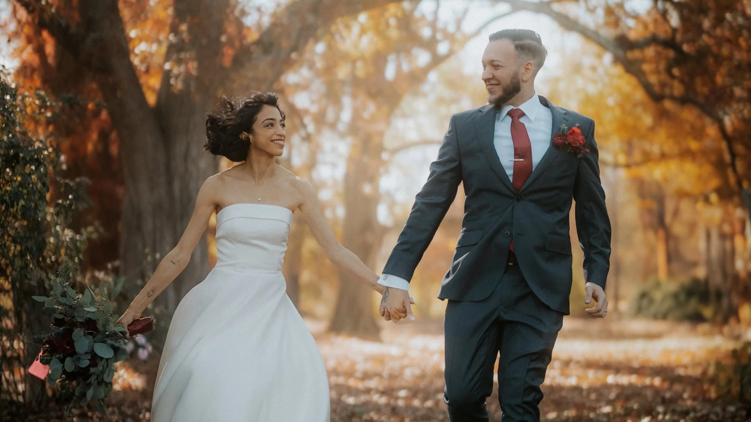 A couple in wedding attire holding hands and walking through an autumn forest, smiling at each other.