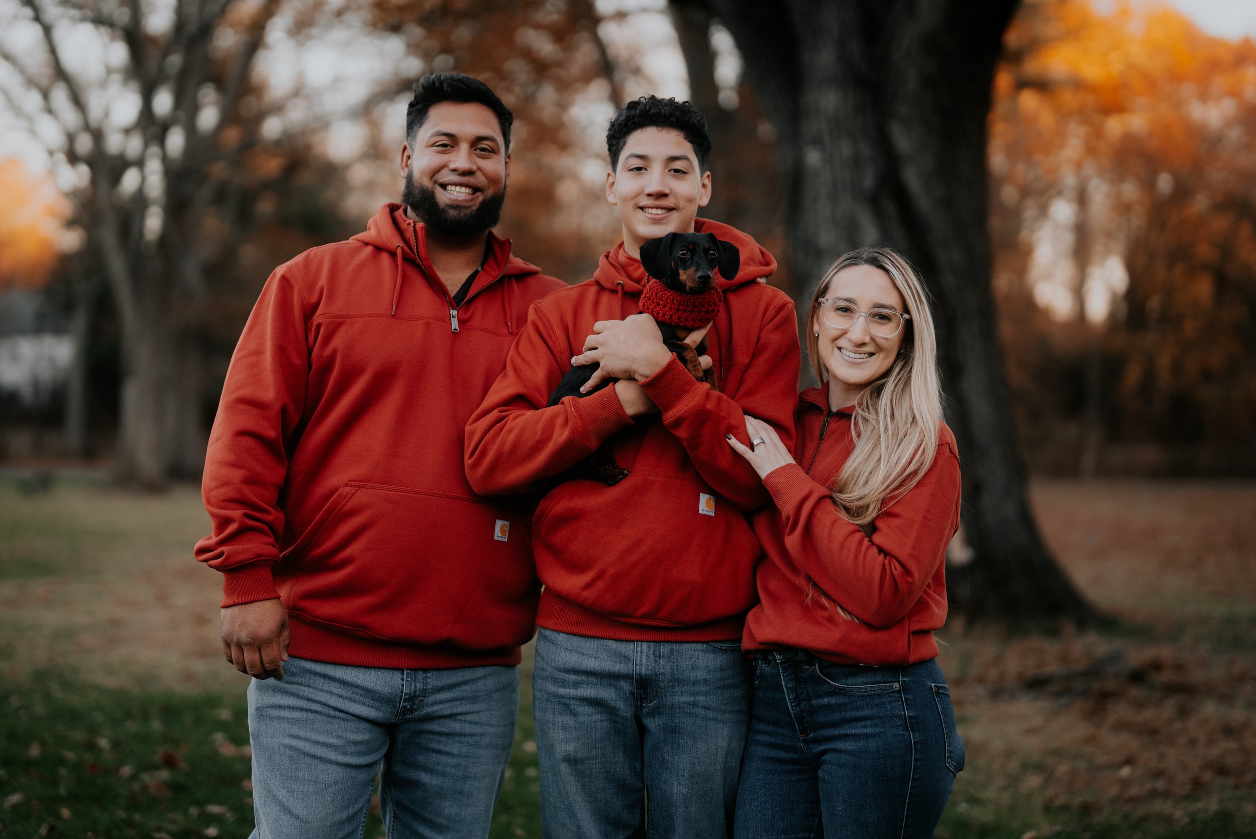 A group of four people and a dog standing outdoors in a park during fall, all wearing matching red hoodies. The man on the left has a beard and is smiling at the camera. The young man in the middle holds a small black dog wearing a red scarf, and the