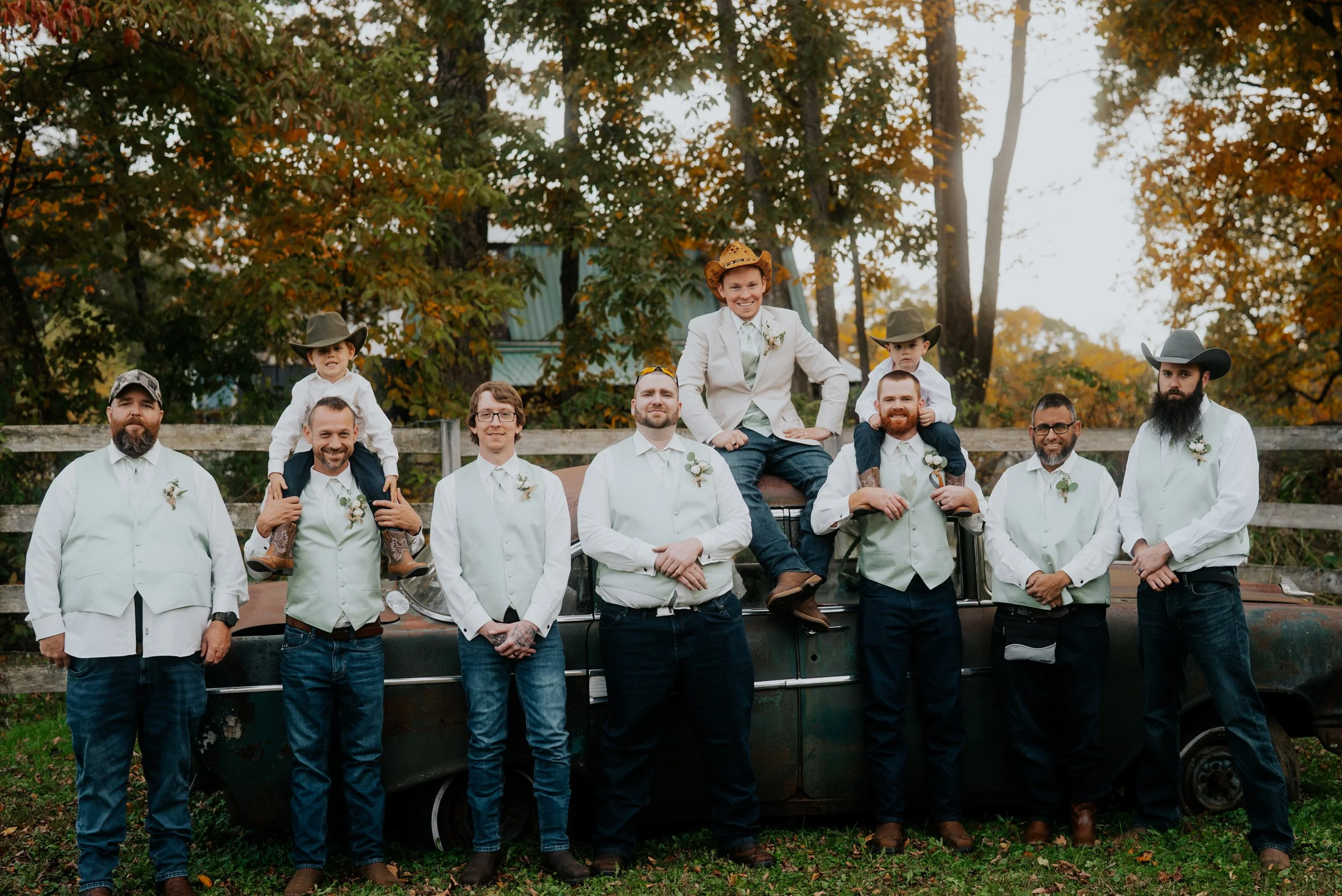A group of eleven men, some with children, posing outdoors in front of a wooden fence and trees with fall foliage. They are dressed in white shirts and vests, with some wearing cowboy hats. The children are sitting on the shoulders of two men, wearin