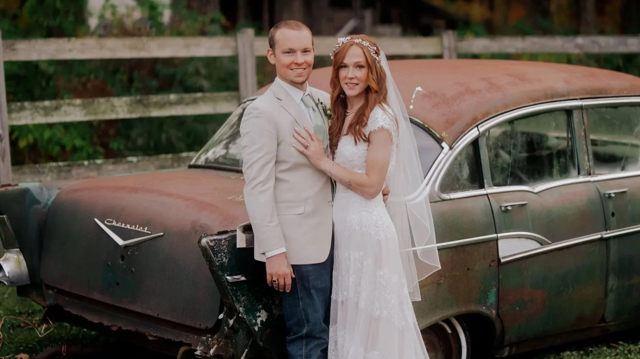 A newlywed couple standing close together in front of an old, rusty Chevrolet car. The groom is wearing a light beige suit jacket with a white shirt and tie, and the bride is in a white lace wedding dress with a veil and floral headpiece. They are smiling and looking at the camera, with a wooden fence and greenery in the background.