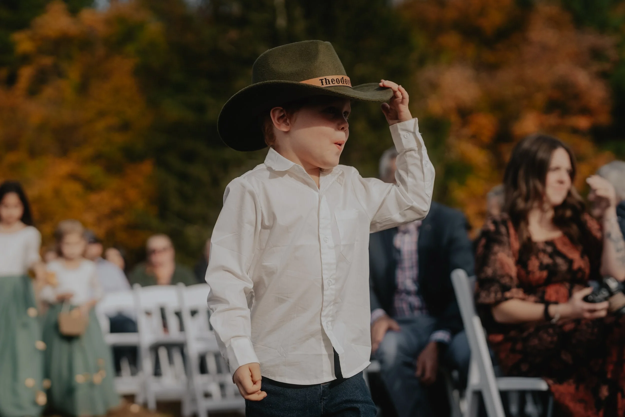 A young boy wearing a white shirt and a large cowboy hat with the name "Theodora" on a band, standing outdoors at what appears to be a gathering or celebration, with people seated in the background against a backdrop of trees with fall foliage.