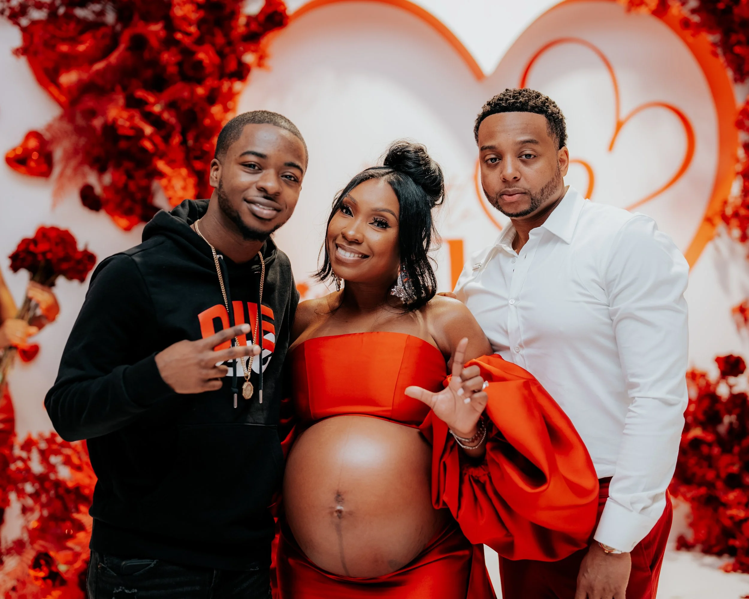 A pregnant woman in a red strapless dress and two men, one in a black hoodie and another in a white shirt, celebrating with her at a romantic Valentine's Day-themed event with red flowers and heart decorations in the background.