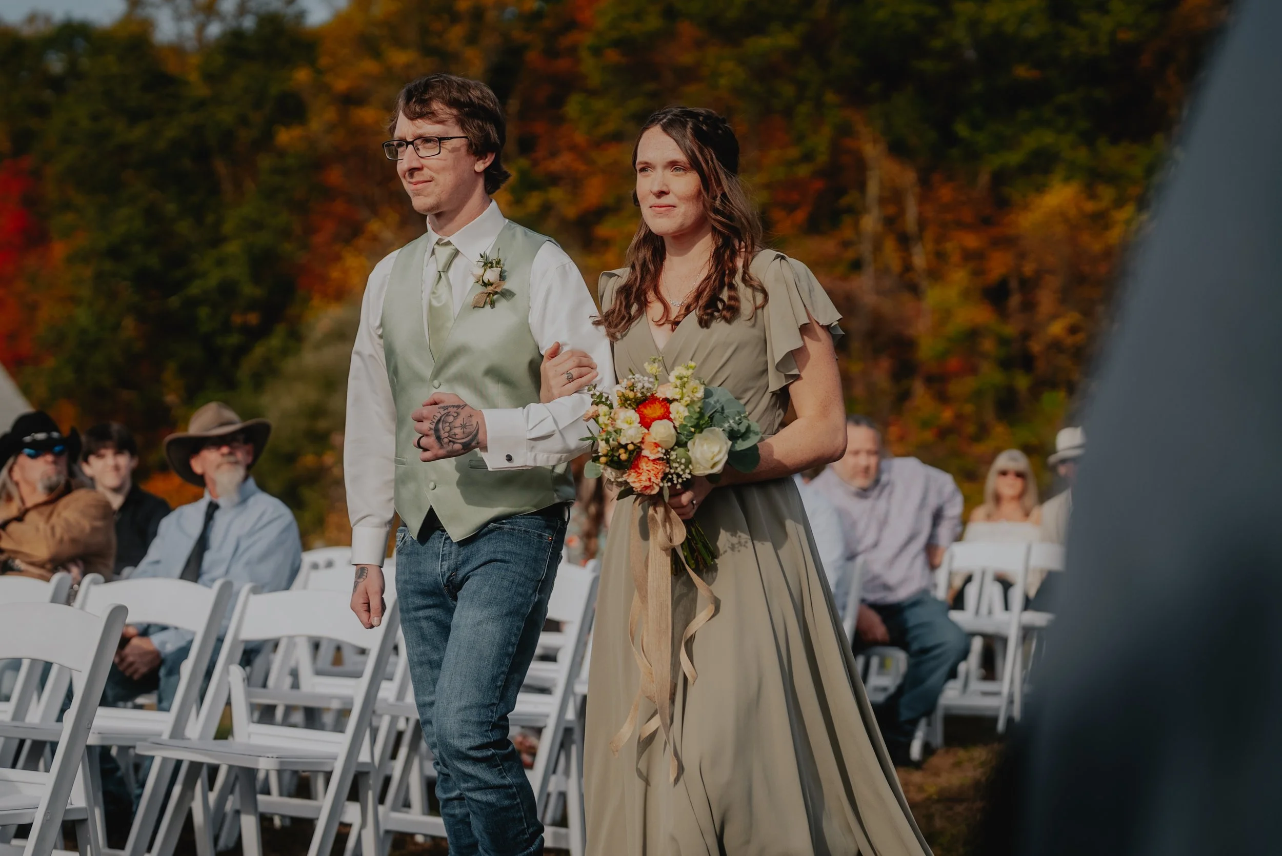 A bride and groom walking down the aisle outdoors during a wedding ceremony, with guests seated on white chairs behind them, autumn trees in the background.