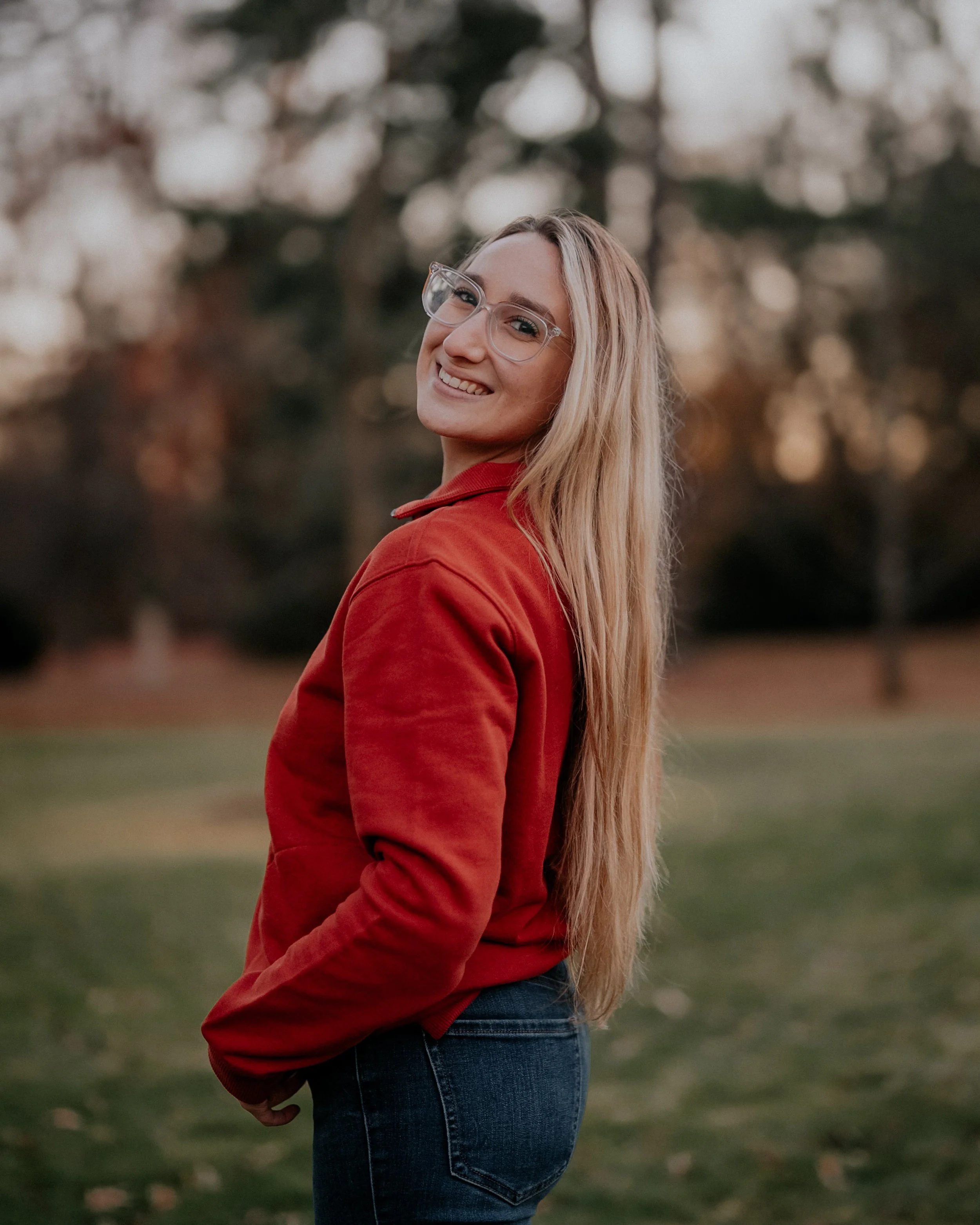 A young woman with long blonde hair, wearing glasses, a red jacket, and jeans, standing outdoors in a park during sunset, smiling at the camera.