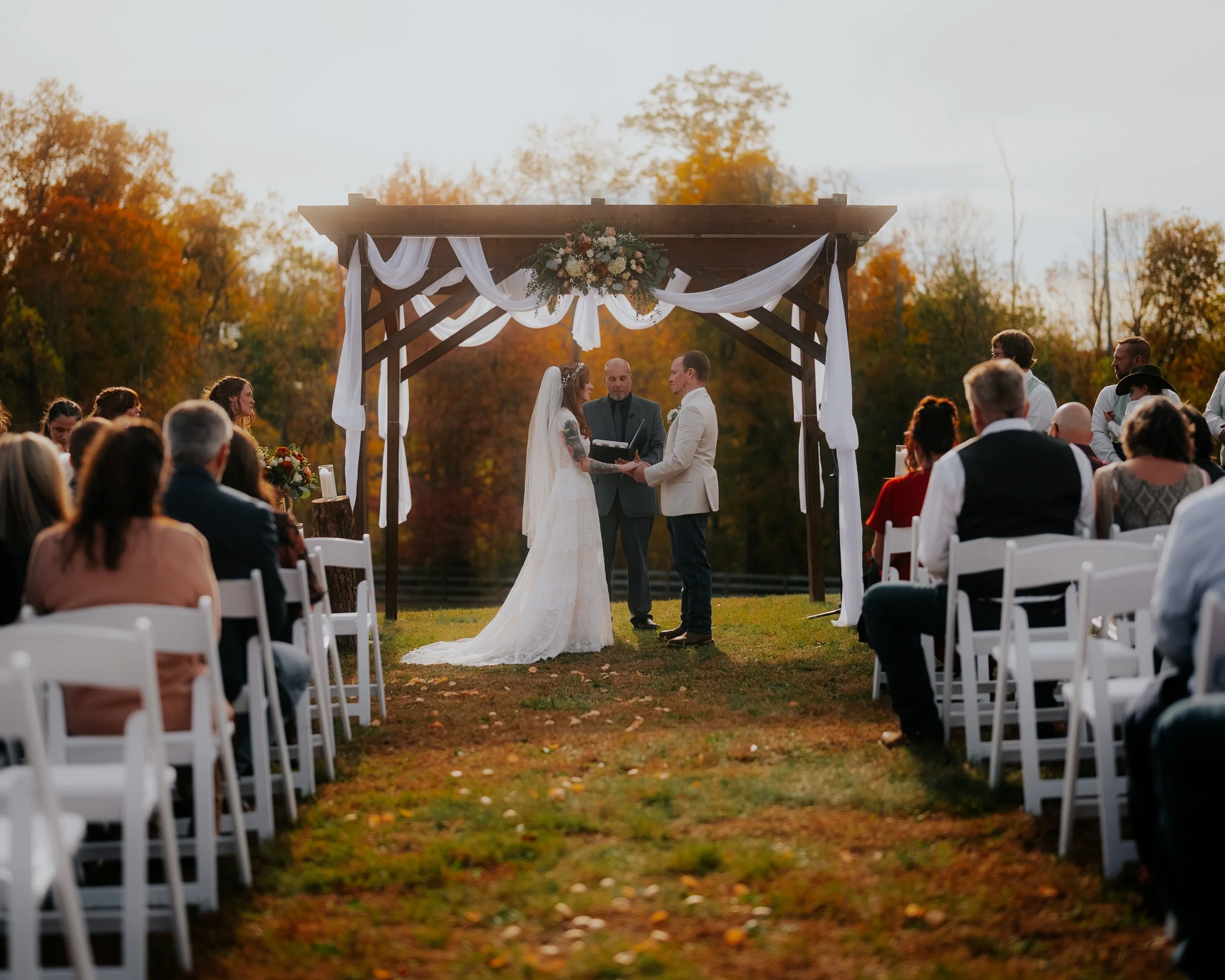 Outdoor wedding ceremony with a bride and groom exchanging vows under a wooden gazebo decorated with white drapery and flowers, surrounded by seated guests with fall foliage in the background.