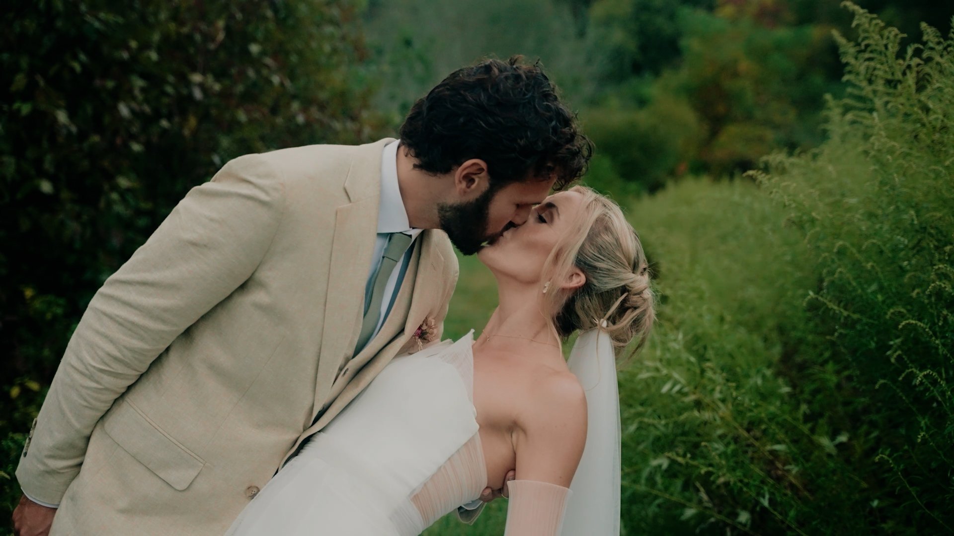 A man and woman in wedding attire sharing a kiss outdoors surrounded by greenery.