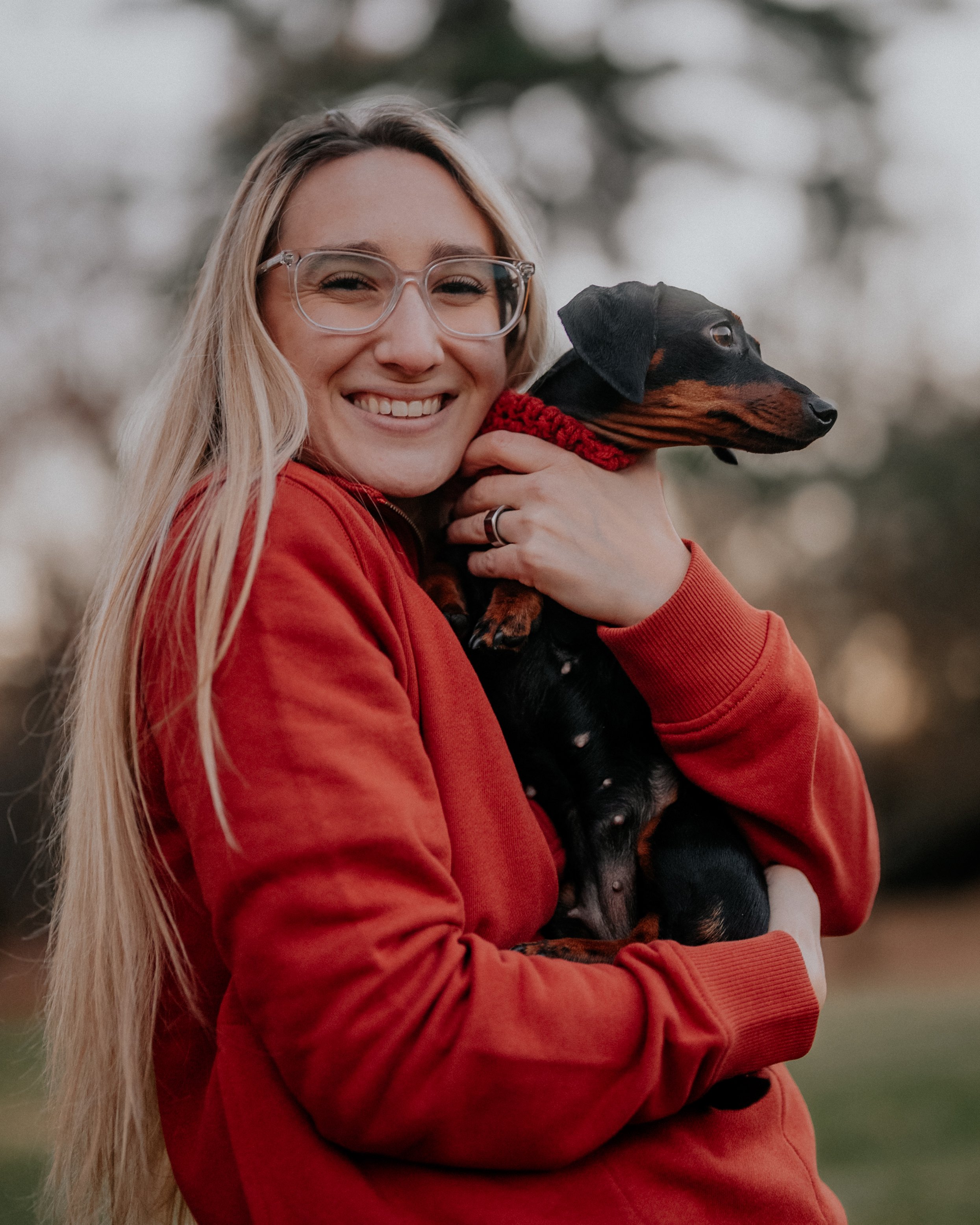 A woman with long blonde hair, glasses, and a red hoodie, smiling while holding a small black and tan dachshund dog with a red knit collar outdoors.