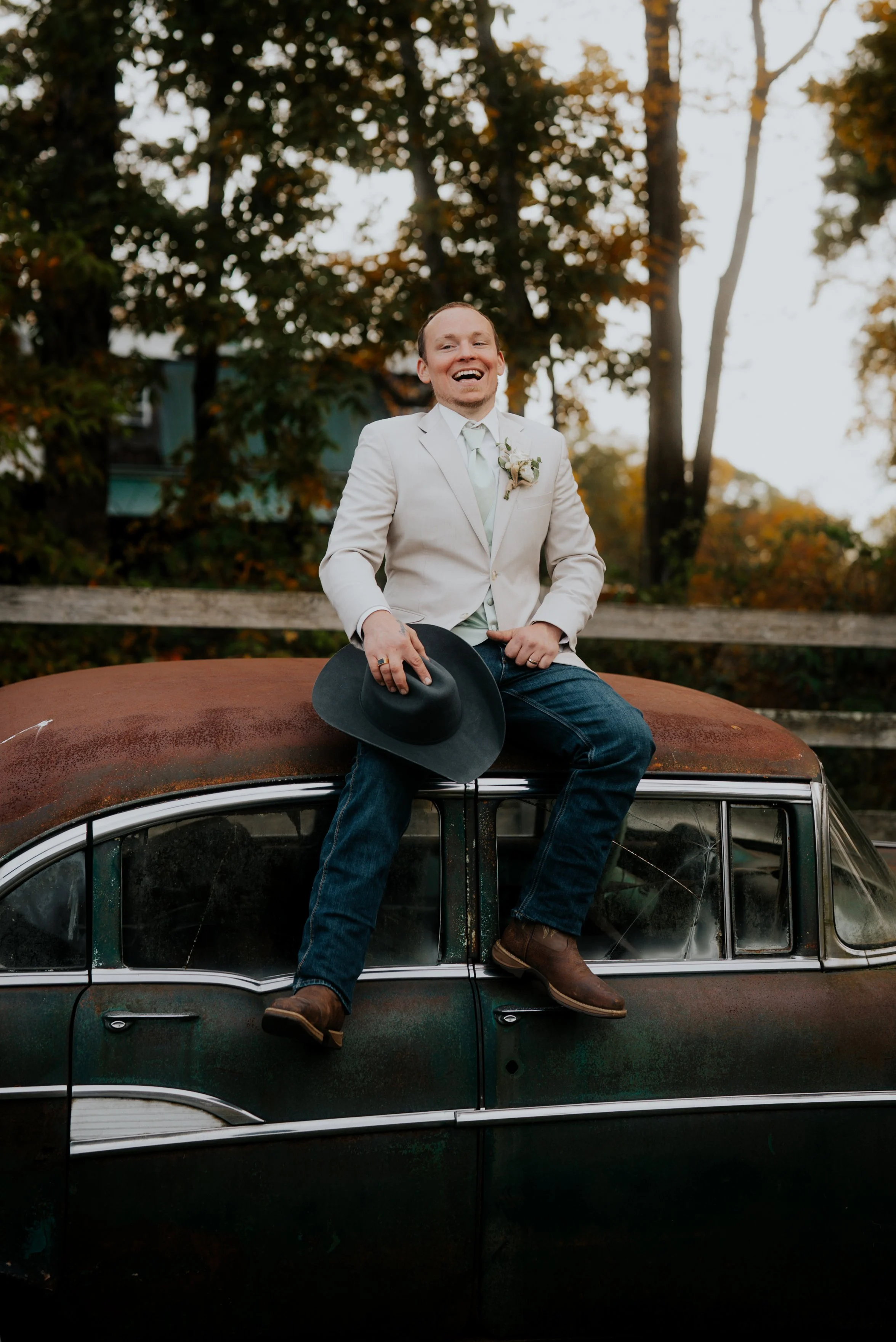A man sitting on the roof of an old rusted car outdoors, holding a wide-brimmed hat, smiling, wearing a white blazer, light-colored shirt, jeans, and brown boots, with trees and a rustic wooden fence in the background.
