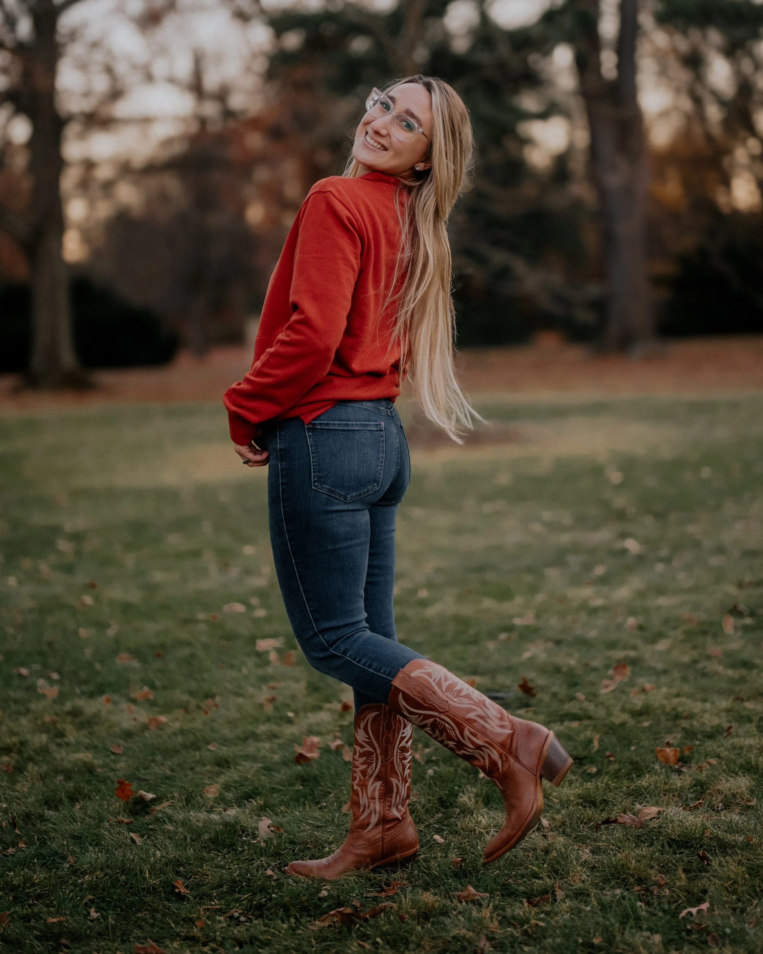 A woman with long blonde hair, glasses, wearing a red sweater, blue jeans, and cowboy boots, standing outdoors on grass with trees in the background, smiling and looking back at the camera.