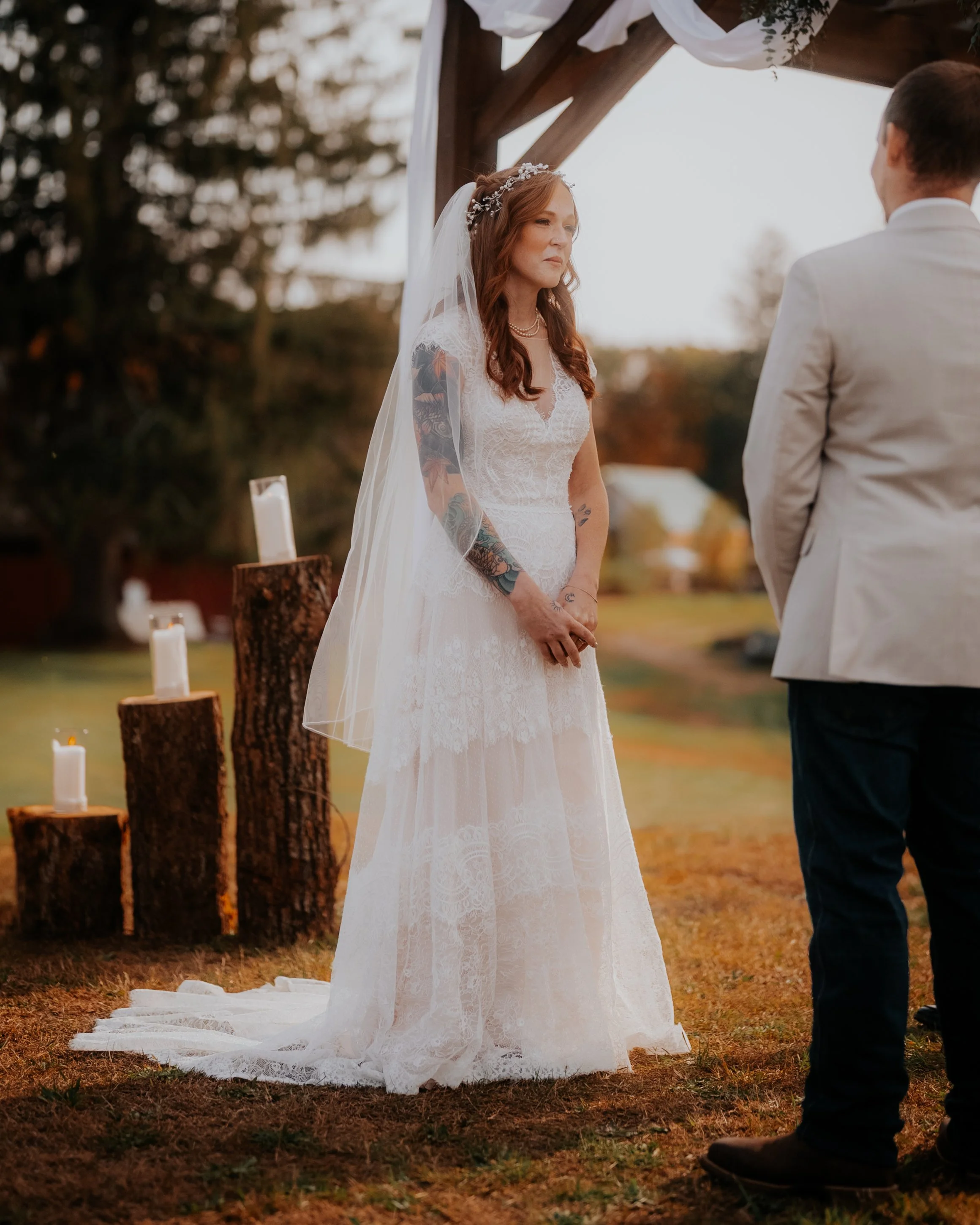 A bride standing during an outdoor wedding ceremony, with the groom facing her, under a wooden arch decorated with white fabric, in a natural setting with trees and candles on tree stumps.