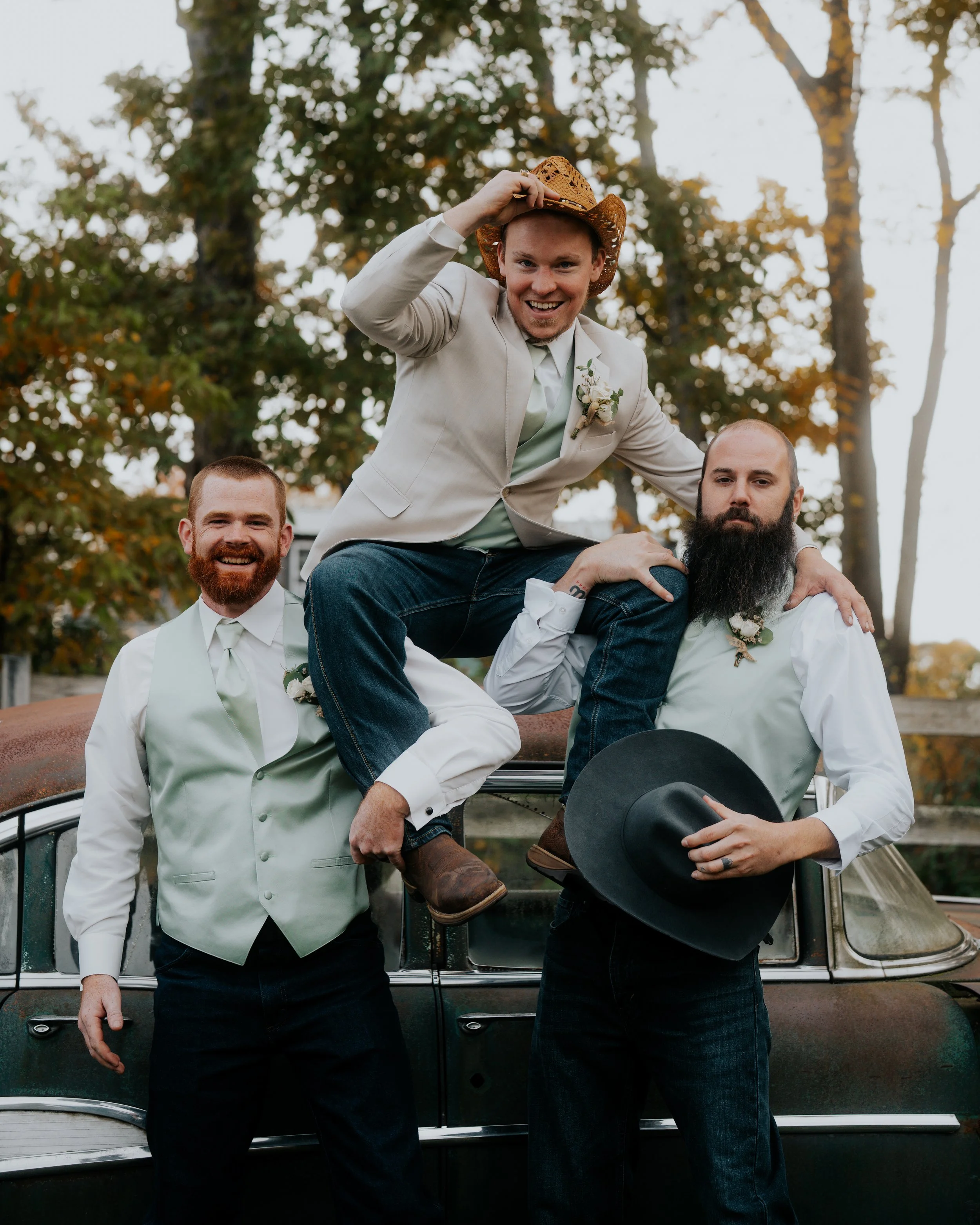 A group of four men, dressed in semi-formal attire, posing outdoors on and around a vintage car with trees and fall foliage in the background. One man is wearing a cowboy hat and leaning on the others, who are smiling and holding a hat.