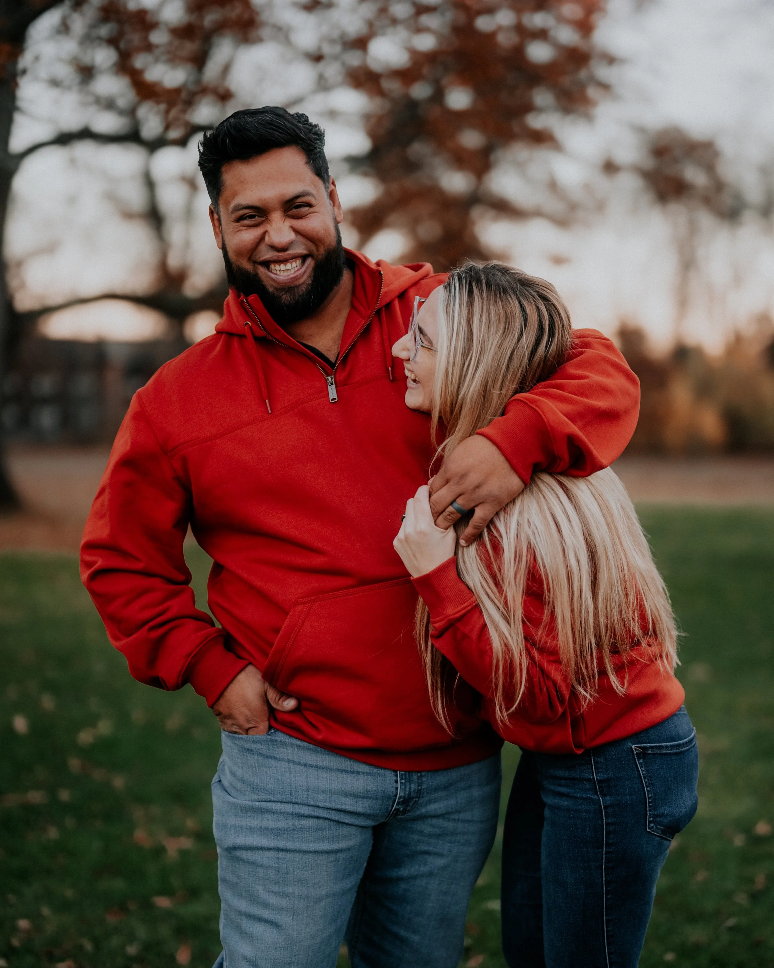 A smiling man with a beard and a woman with glasses sharing a hug outdoors in autumn, both wearing red hoodies.