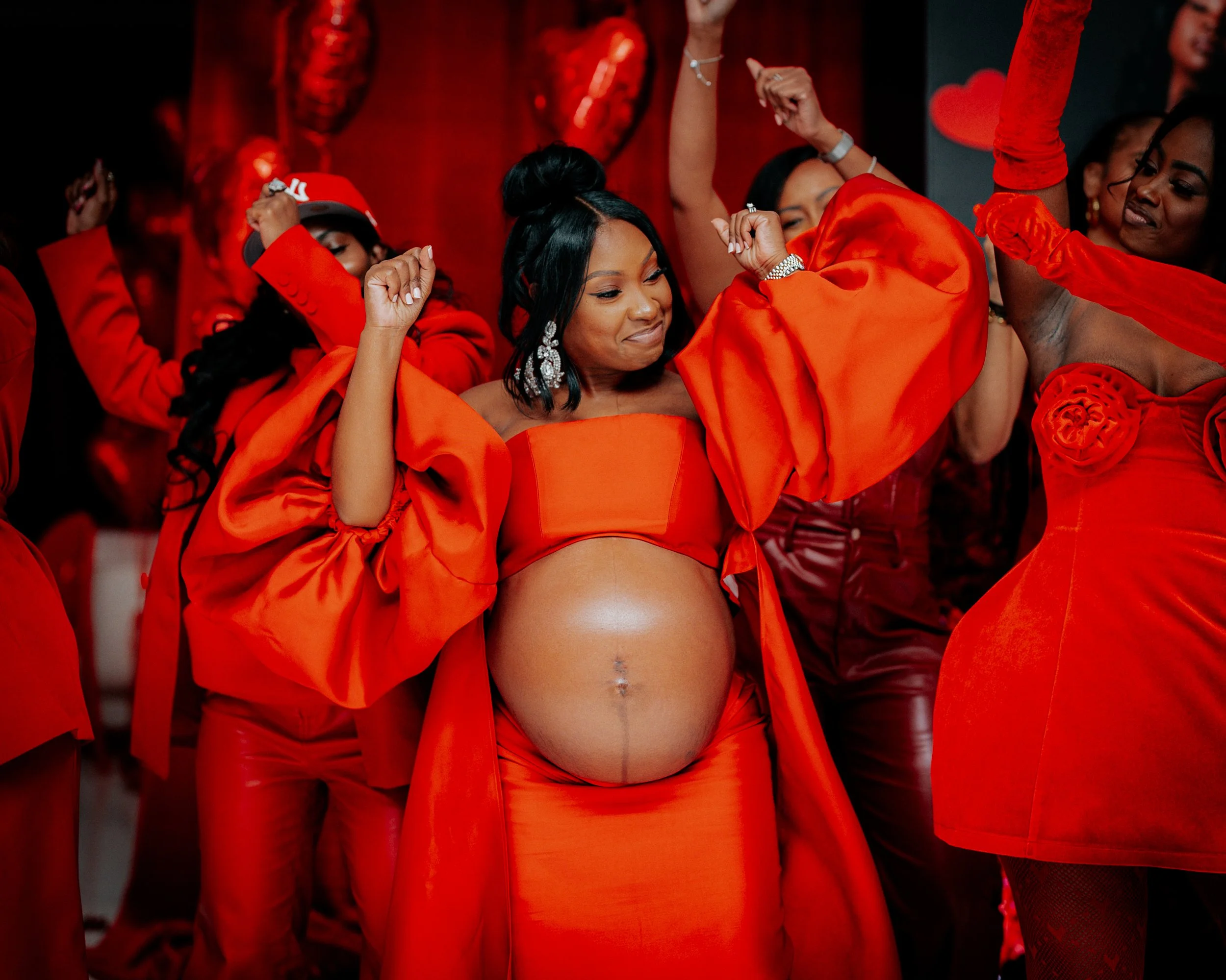 Group of women dancing at a celebration, with one woman in the center exposing her pregnant belly, all dressed in red outfits.