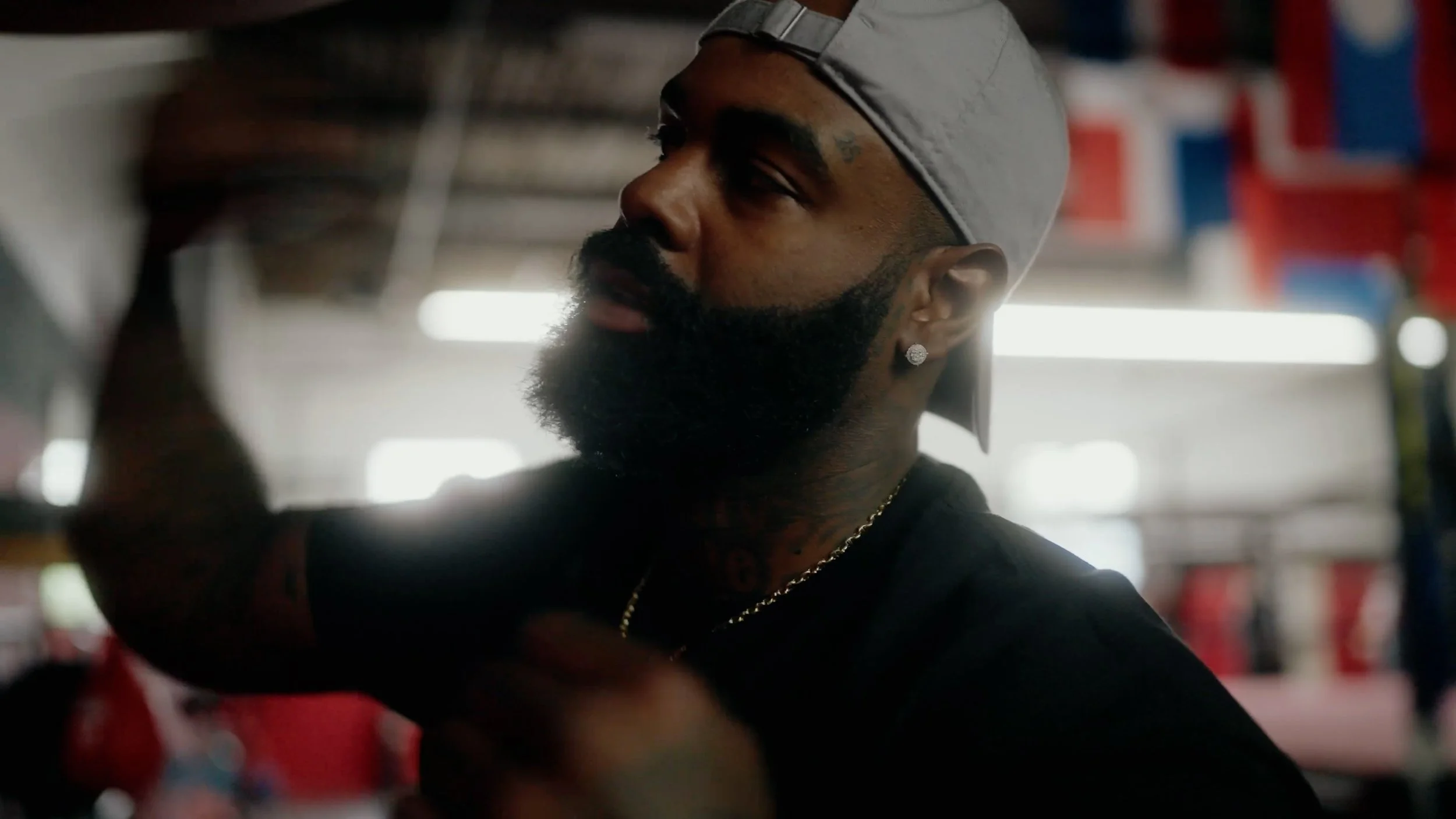 A man with a beard, tattoos, earrings, wearing a gray cap backwards and a black shirt, in a gym or boxing ring, raising his arm in a fist.