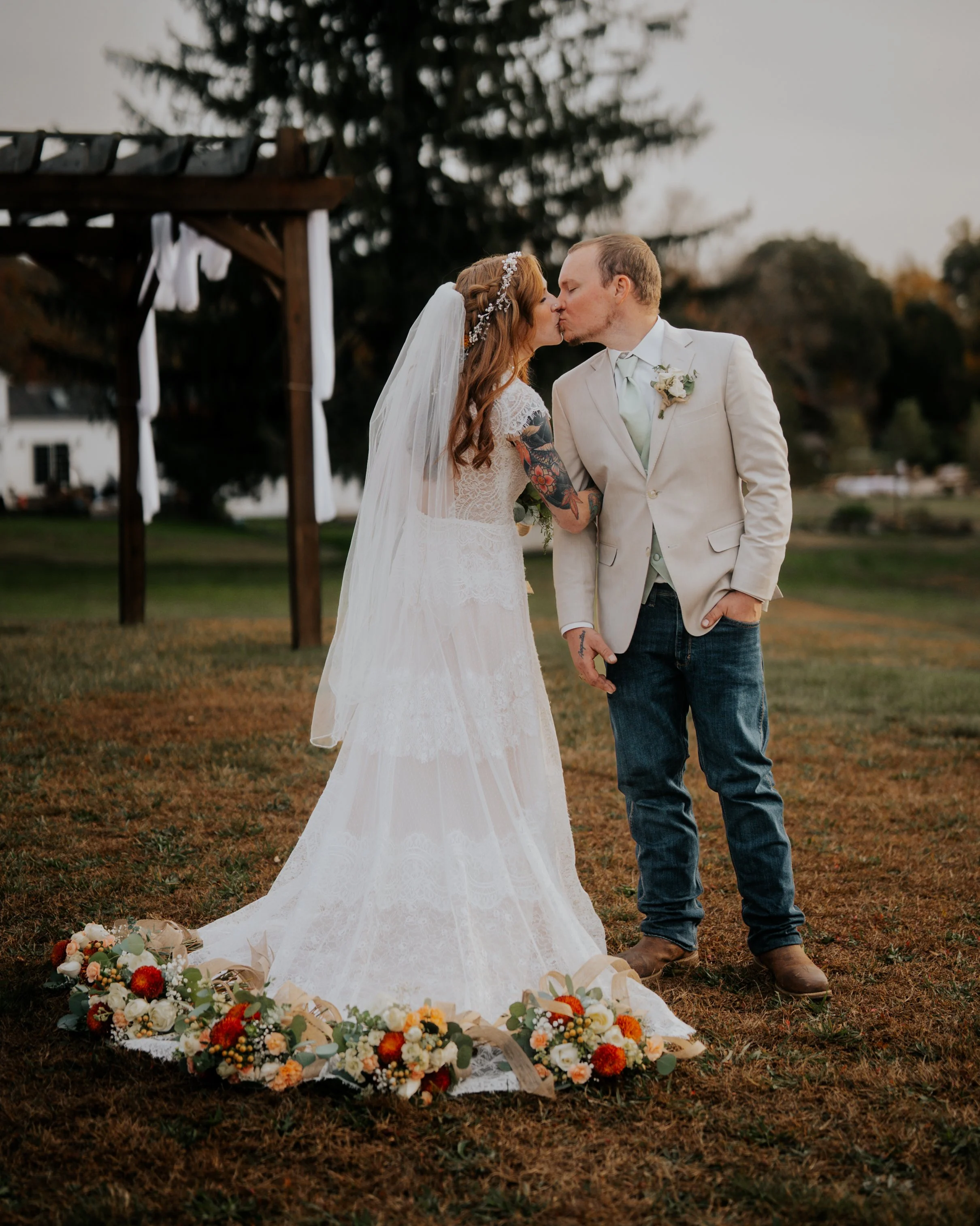 A bride and groom kiss during their outdoor wedding, surrounded by floral arrangements on the ground and a wooden wedding arch in the background.