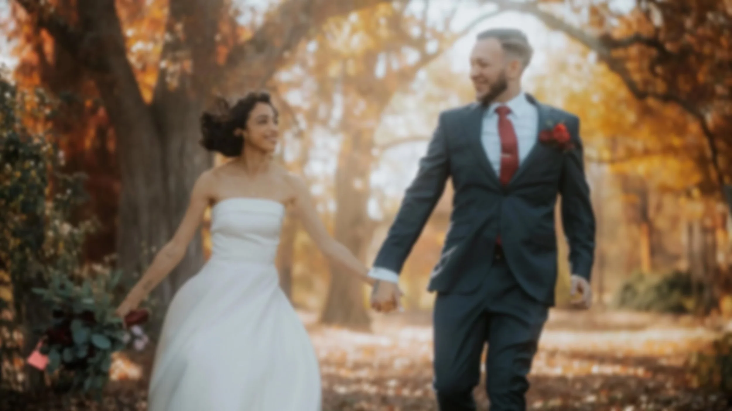 A bride and groom holding hands and running through a park with autumn trees and fallen leaves.