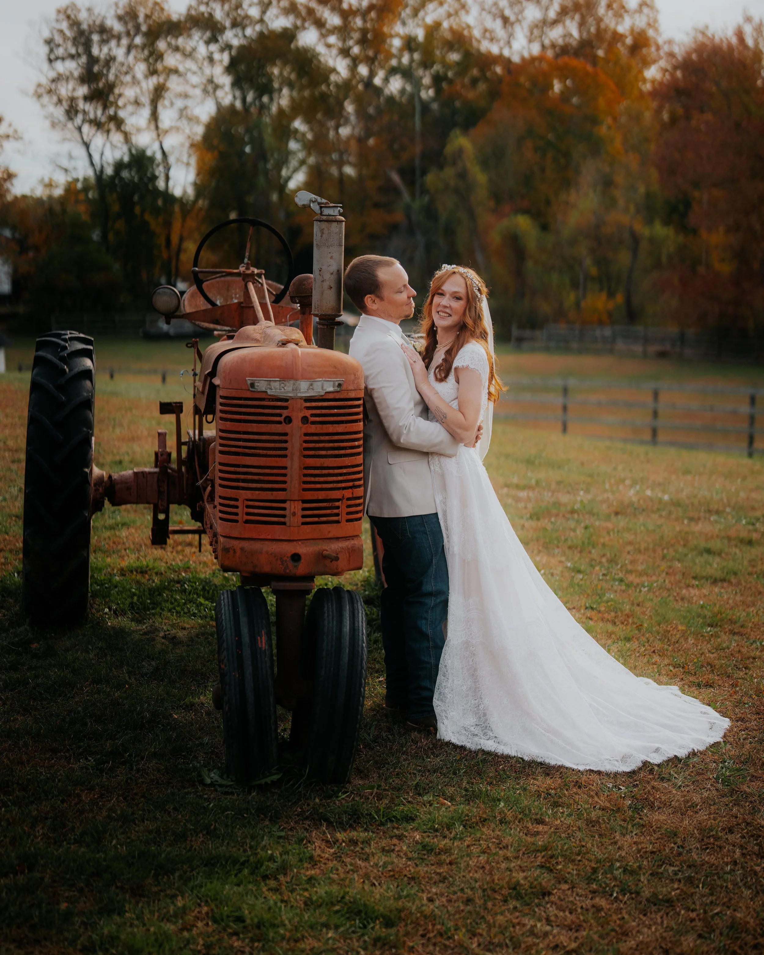 A couple dressed in wedding attire, the woman in a white wedding dress and the man in a light-colored jacket, embrace and smile while standing next to an old tractor on a grassy field during sunset, with trees in the background.