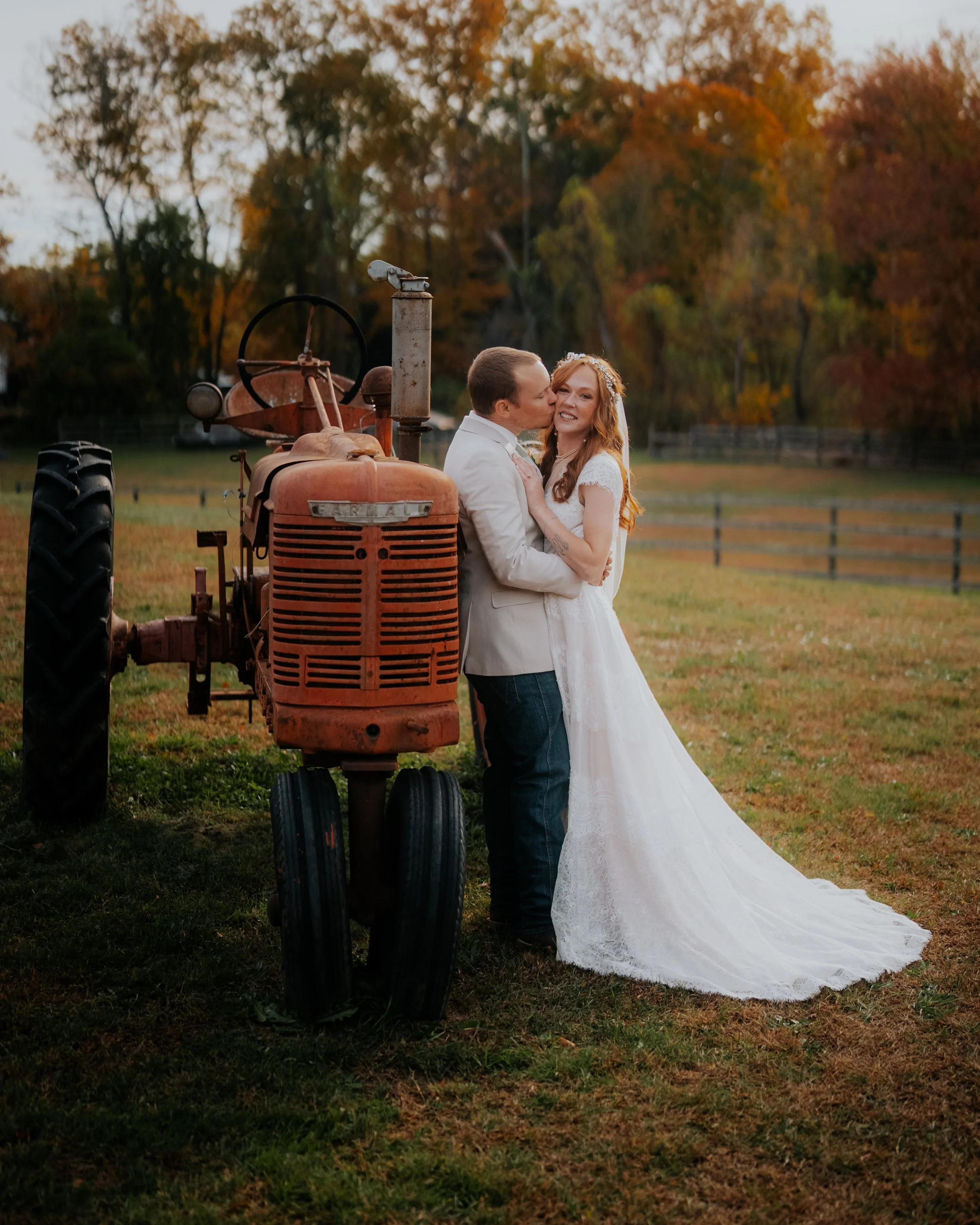 A newlywed couple in wedding attire sharing a kiss and embracing in a field next to an old rusted tractor, with autumn trees in the background during sunset.