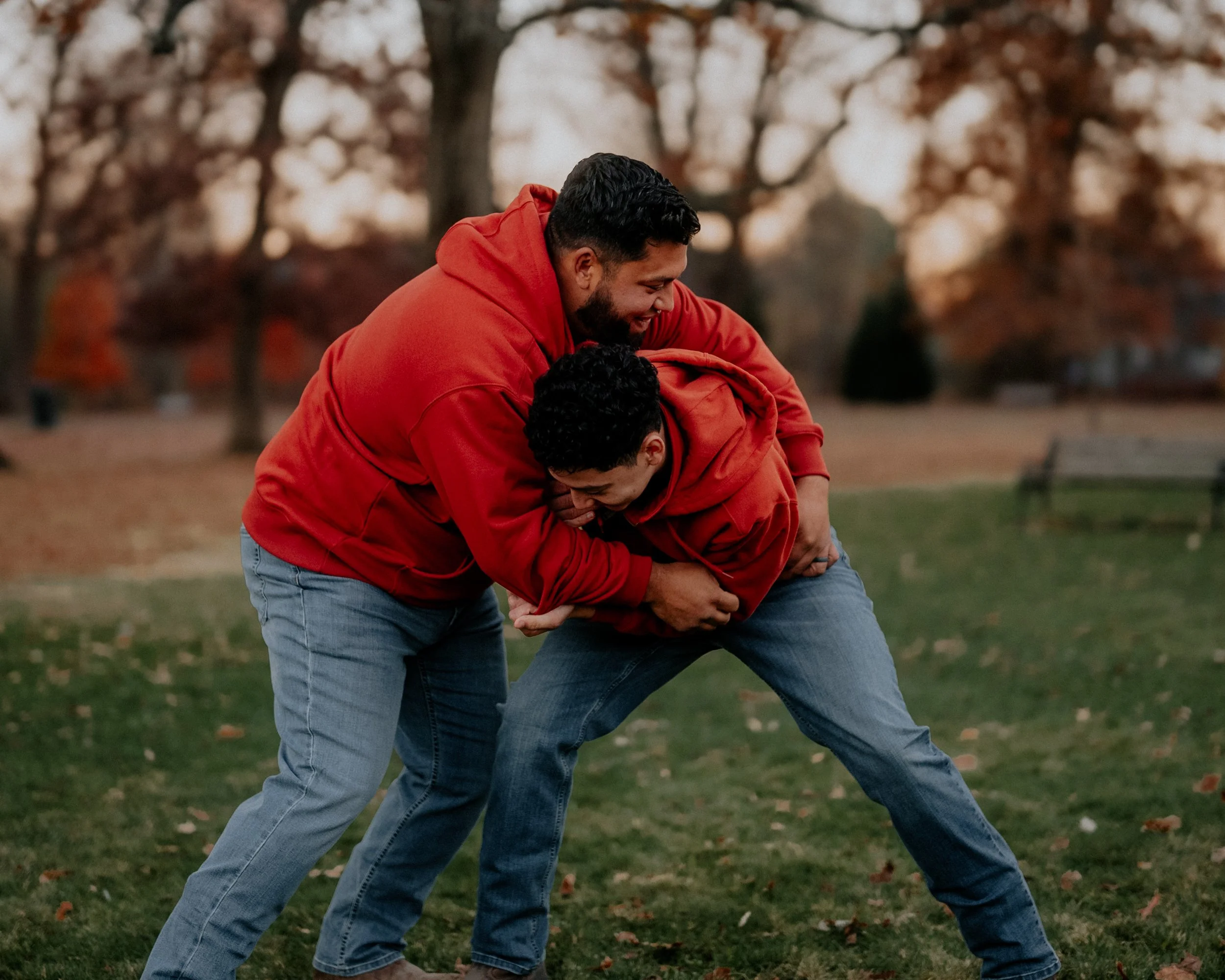 Two men are engaged in a playful wrestling match outdoors in a park, wearing red hoodies and jeans, with trees and fallen leaves in the background.