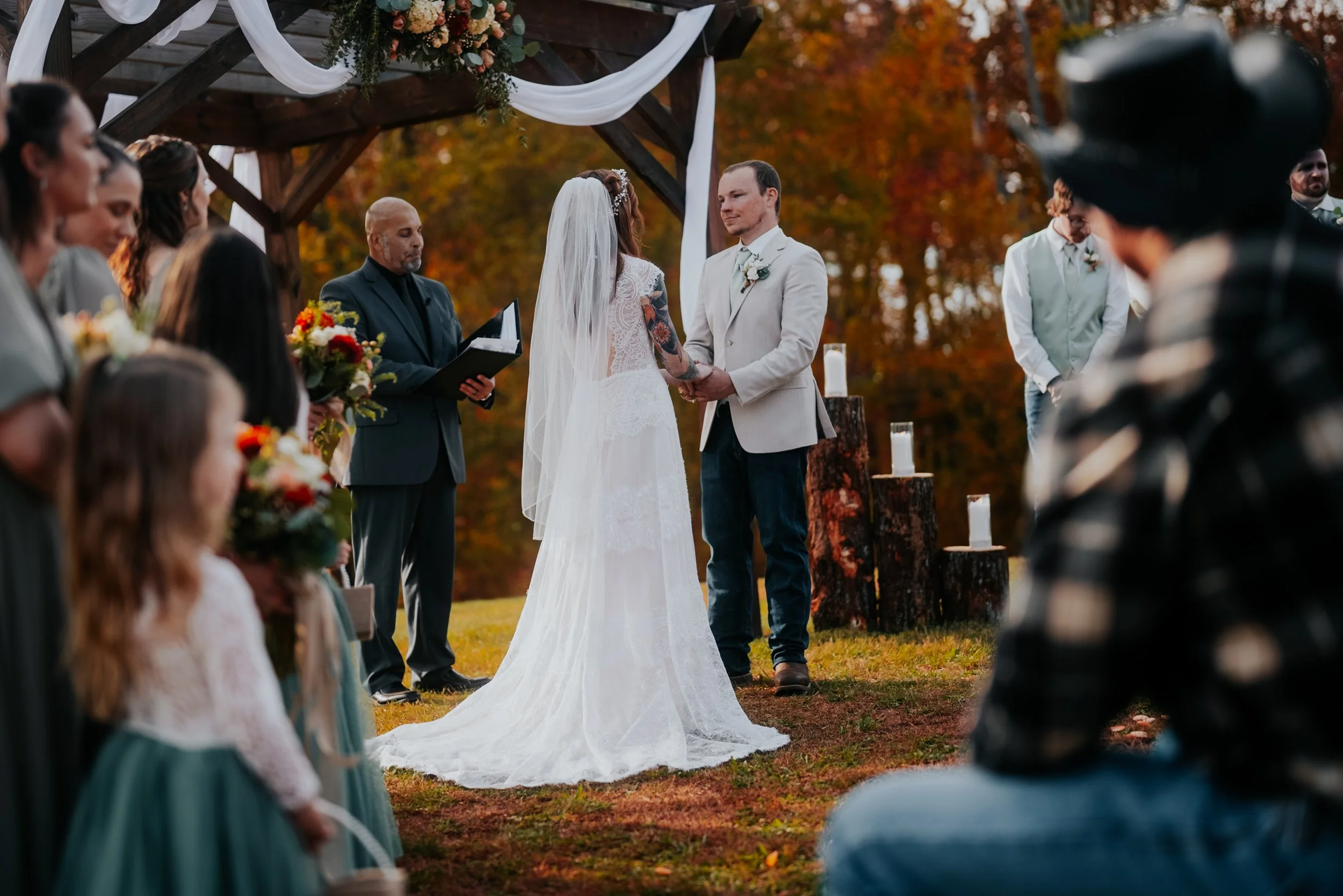 A wedding ceremony outdoors in autumn, with the bride and groom holding hands at the altar, officiant reading, guests watching, and fall trees in the background.