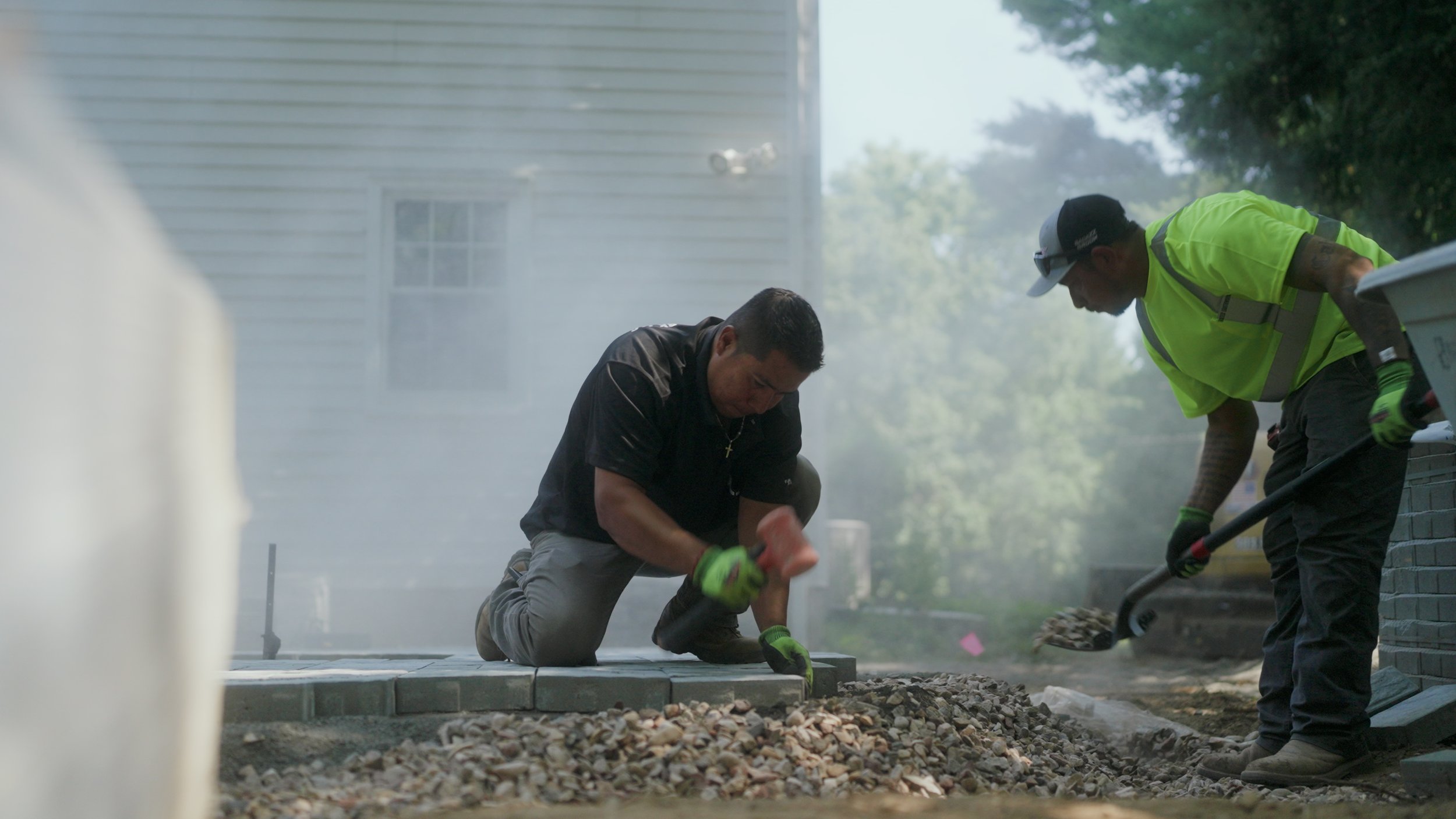Two men working on a construction site, one kneeling down and placing bricks, the other using a tool on gravel, with a building and trees in the background.
