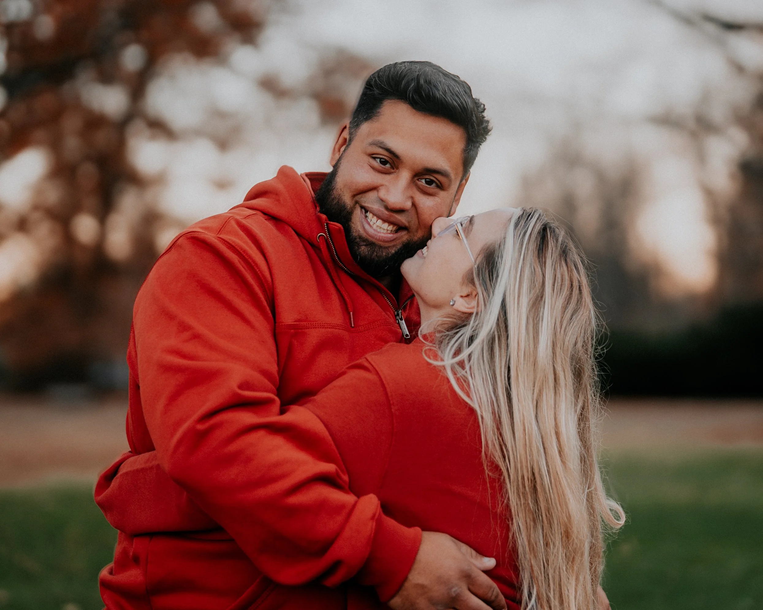 A couple sharing a hug outdoors, both wearing red hoodies, with trees in the background during sunset.