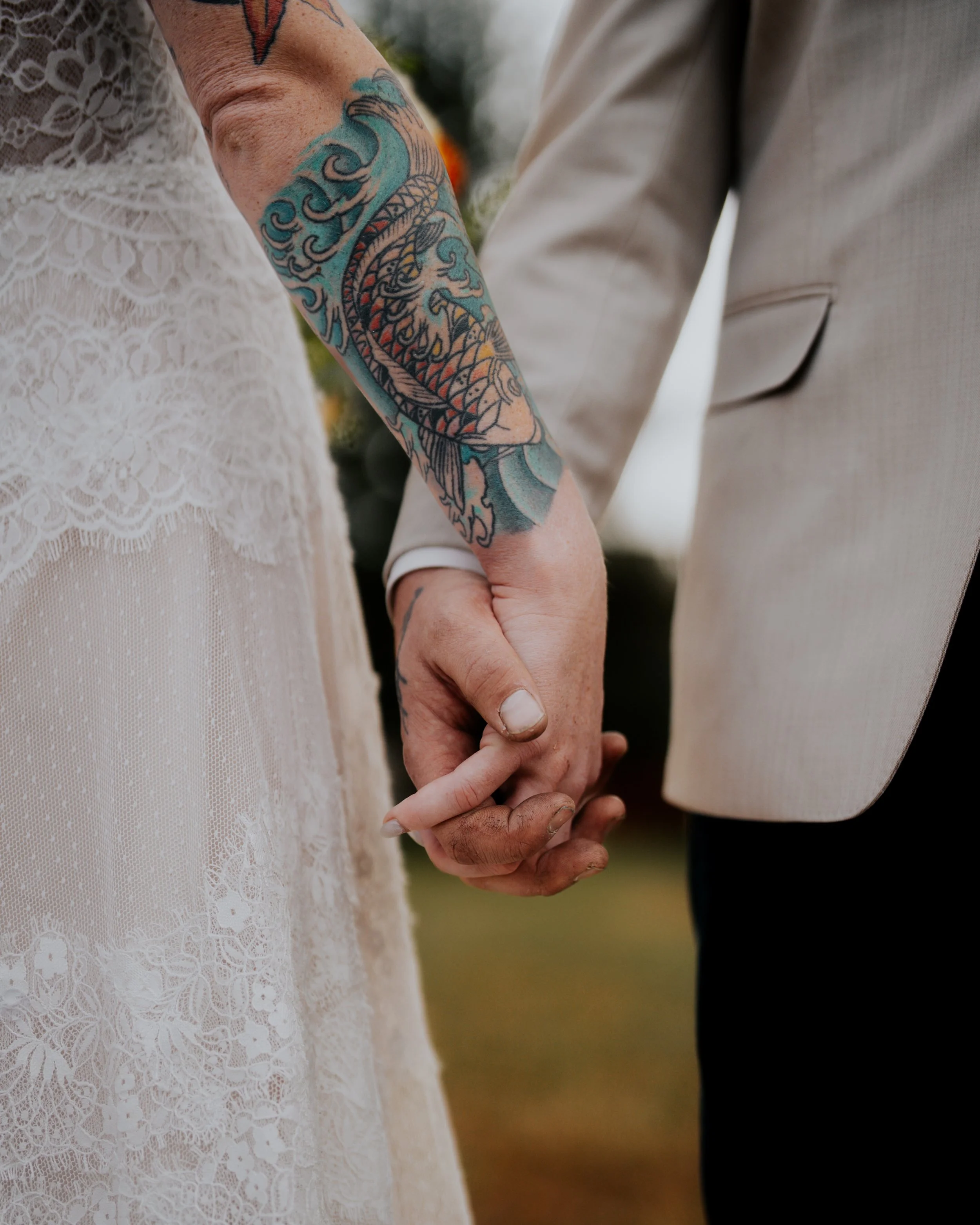 Close-up of a couple holding hands during a wedding, with the woman's lace dress and a colorful tattooed arm visible.