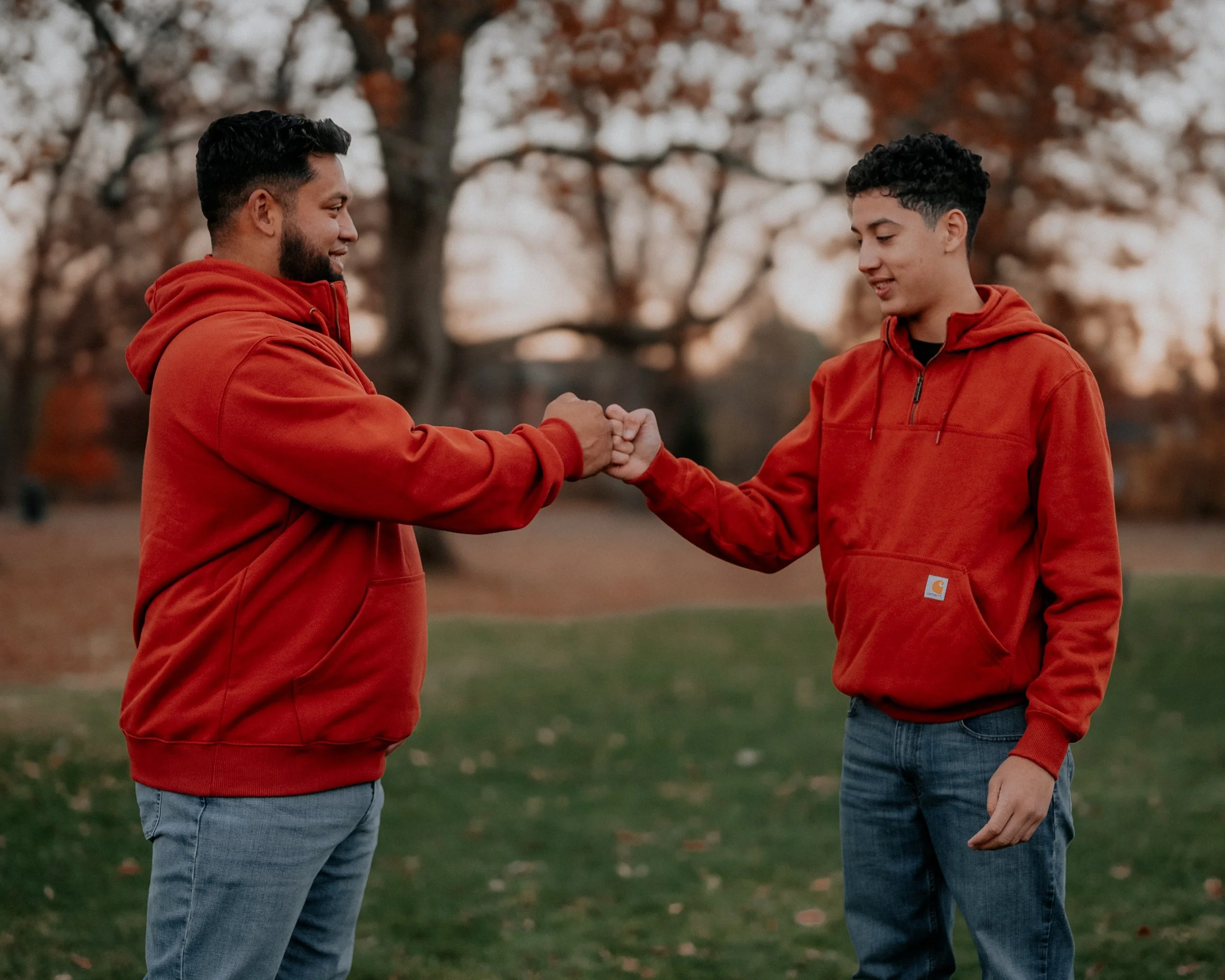 Two males in red hoodies fist bumping outdoors in a park during autumn.