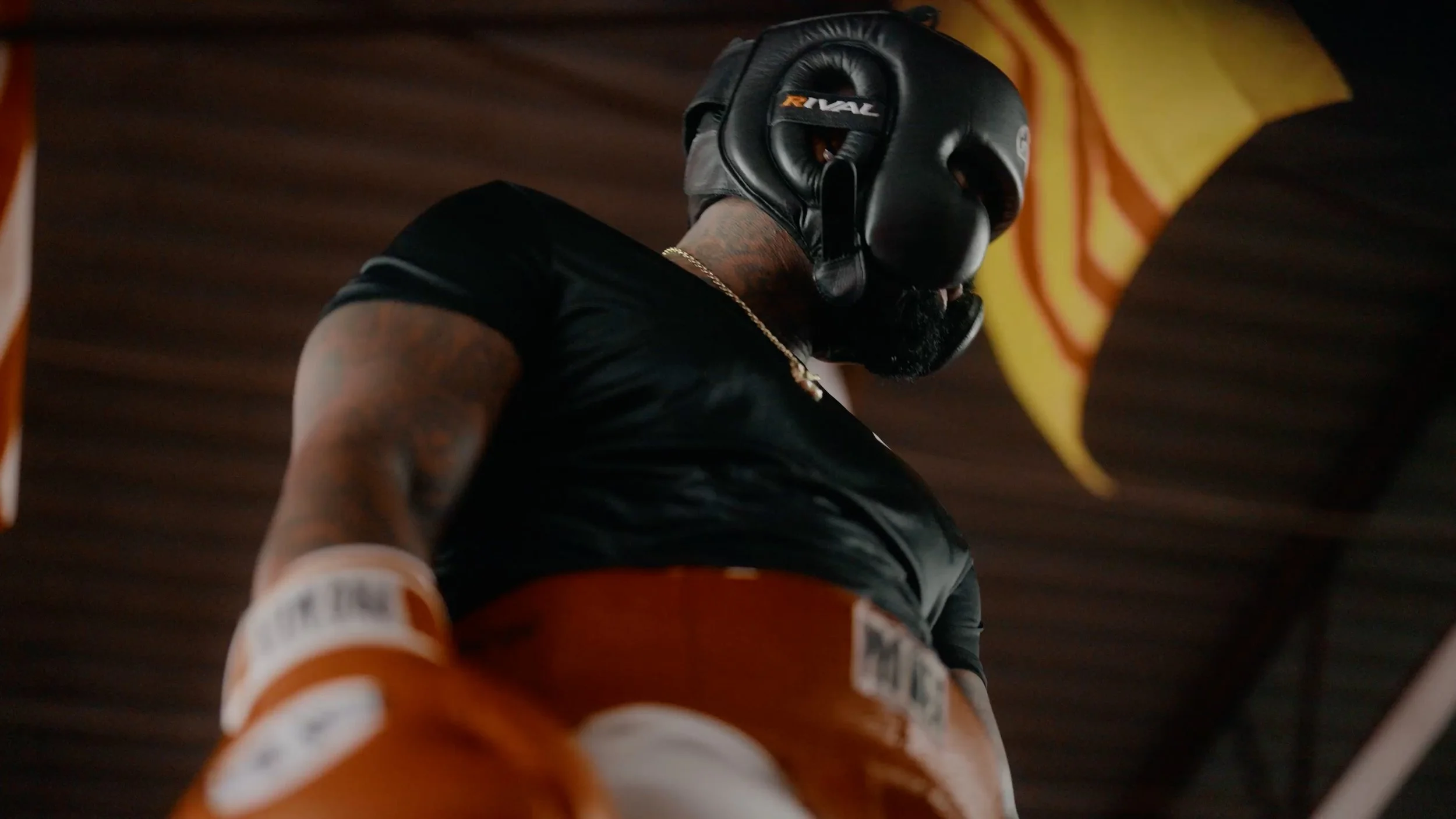 A close-up of a boxer wearing a black headgear, black t-shirt, orange boxing gloves, and a gold chain, with a wooden ceiling and yellow-orange flags in the background.