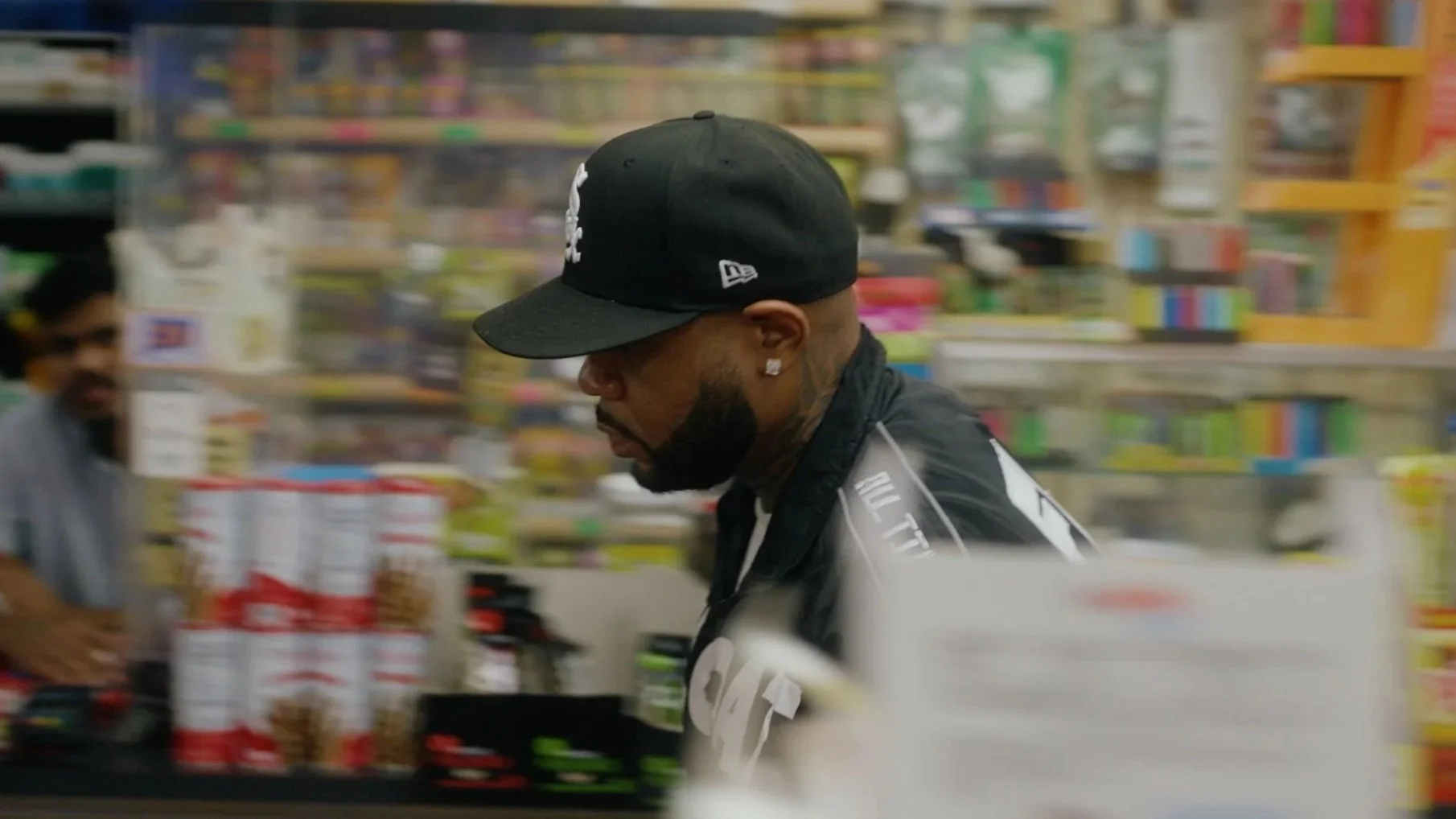A man shopping at a grocery store, wearing a black cap and black shirt, with shelves of colorful products in the background.