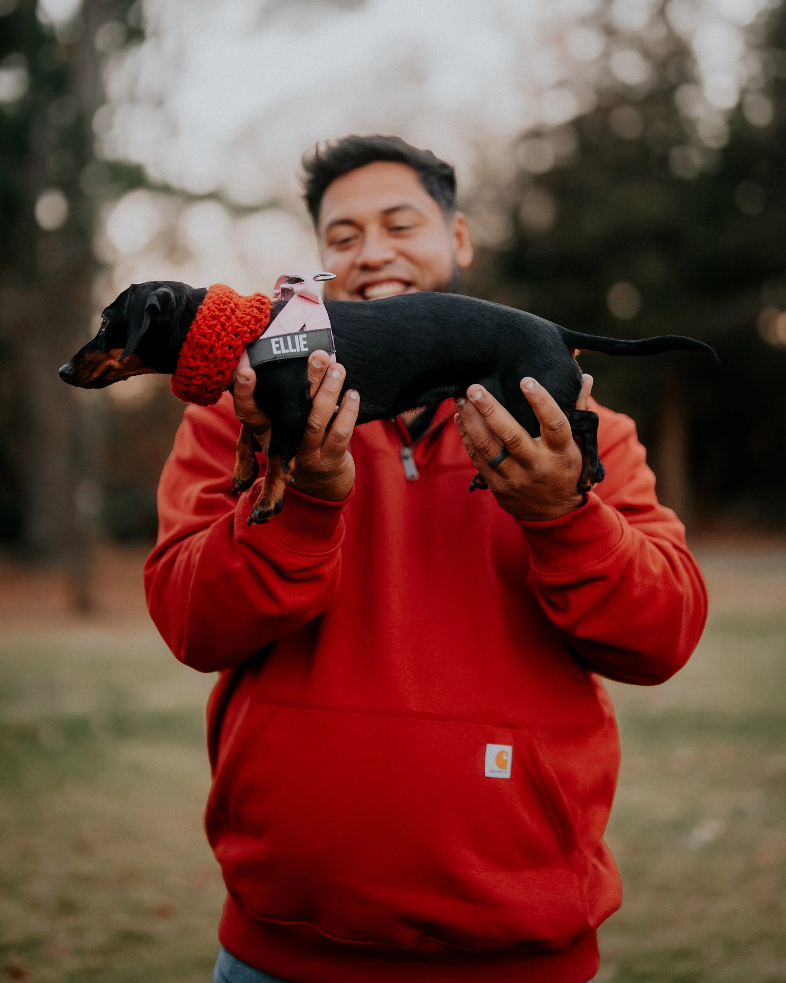 A man in a red hoodie holding a black and tan miniature dachshund wearing a red coat outdoors with trees in the background.