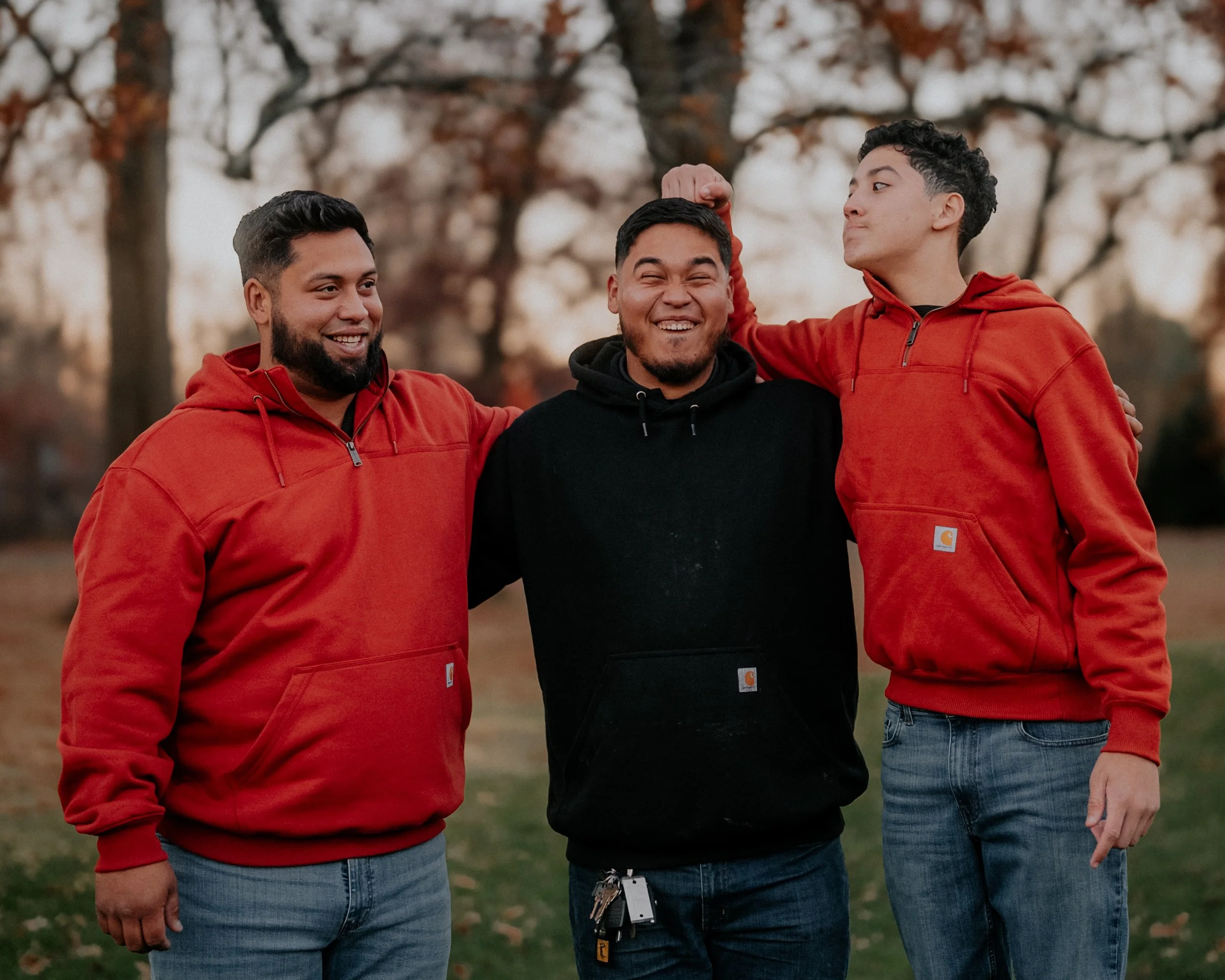 Three young men standing outdoors in autumn, smiling and with arms around each other. One man wears a black hoodie, the other two wear red hoodies.