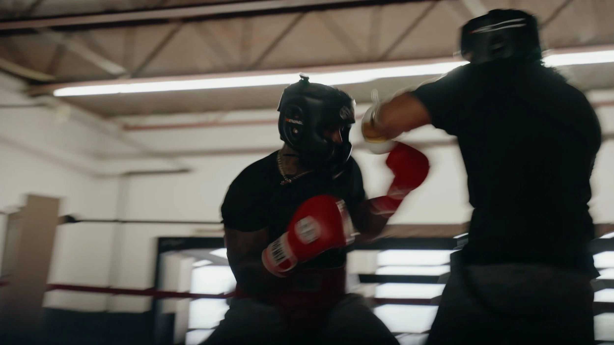 Two men practice boxing in a gym, one wearing a helmet and red boxing gloves, the other in black clothing, throwing punches.