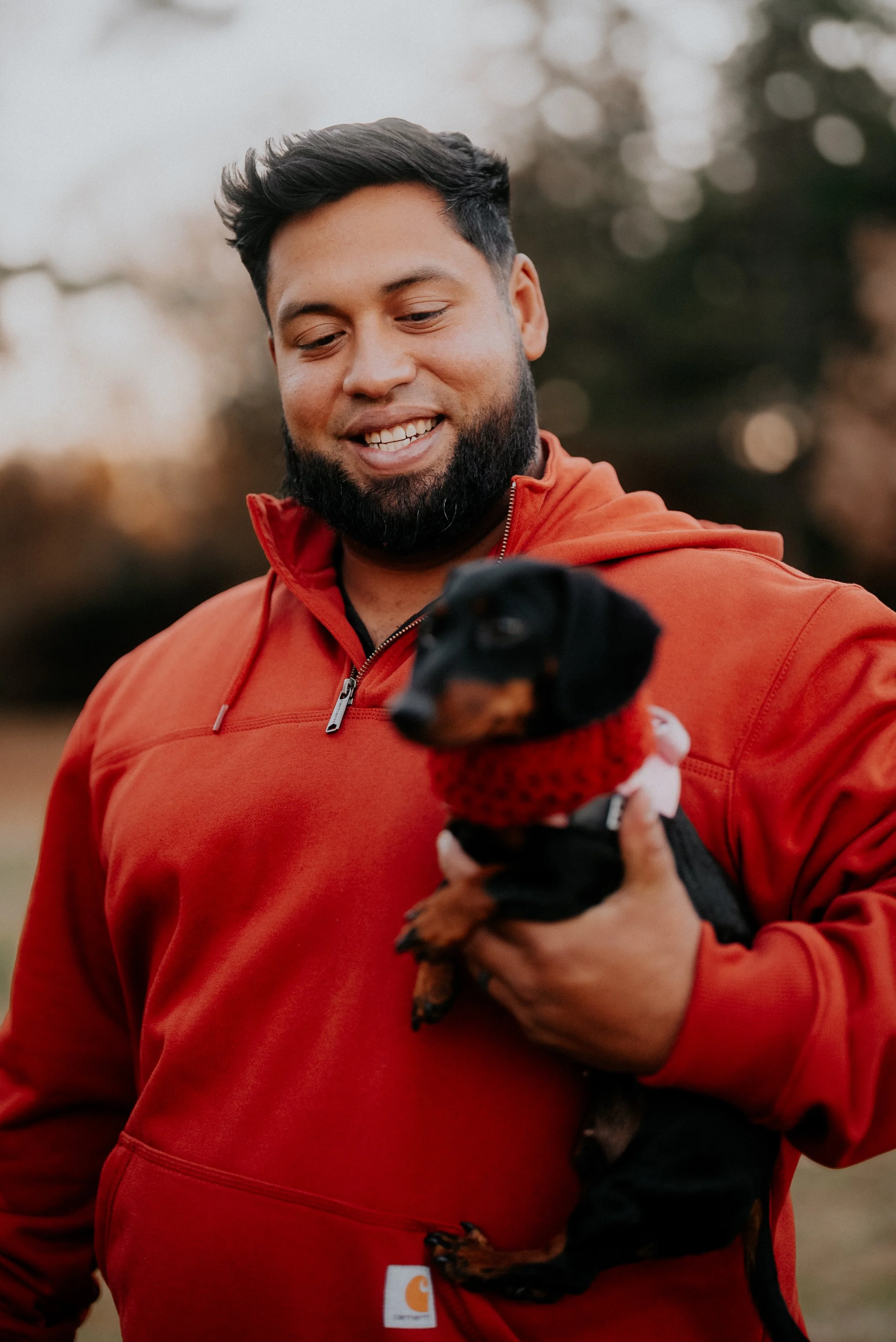 A man with dark hair and a beard is holding a small black and tan puppy wearing a red sweater outdoors at sunset or dusk.