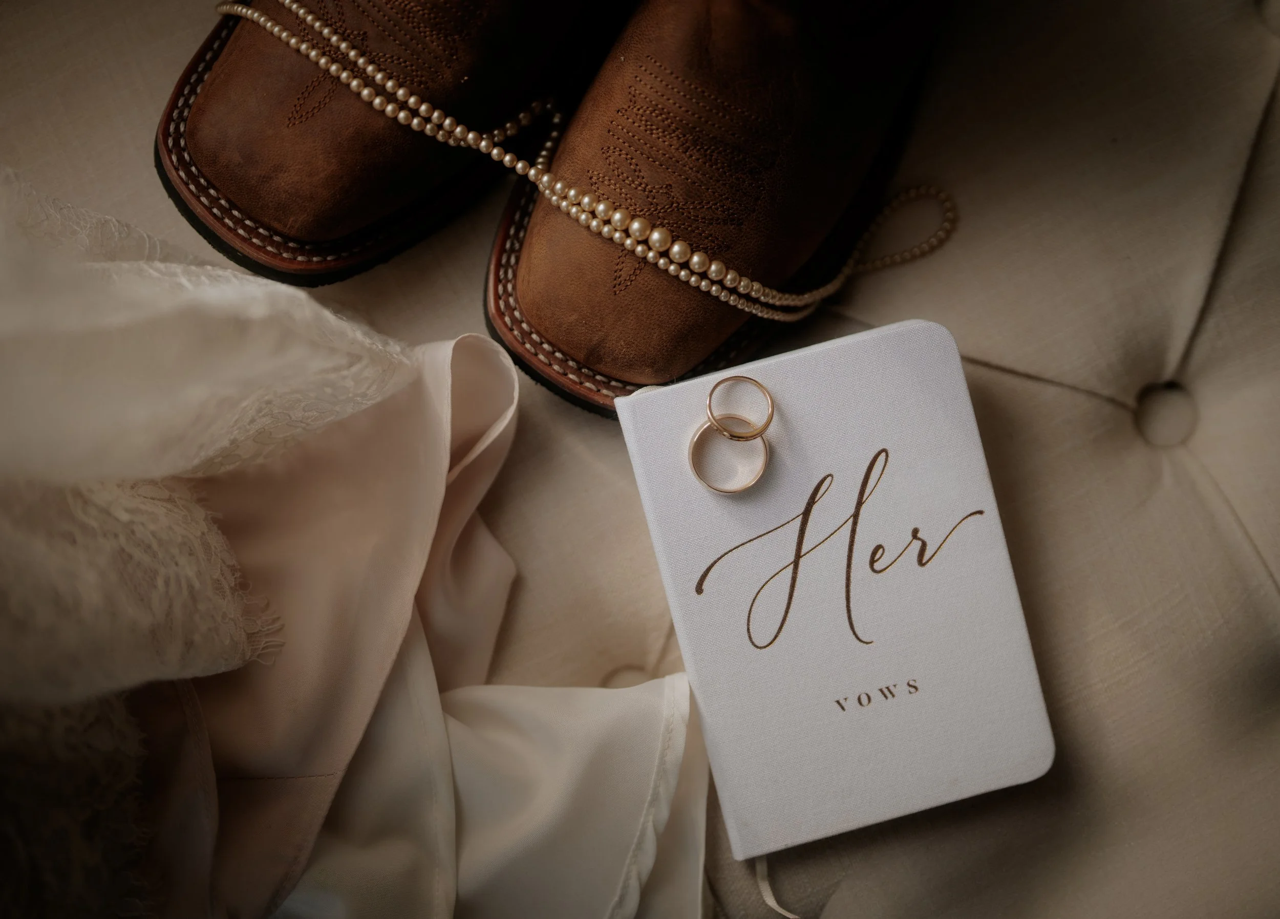 A pair of brown cowboy boots with pearl embellishments, a string of pearls, a beige silky fabric, two gold rings, and a white card with the word 'Her' written in cursive and 'VOUS' underneath, all arranged on a beige quilted surface.