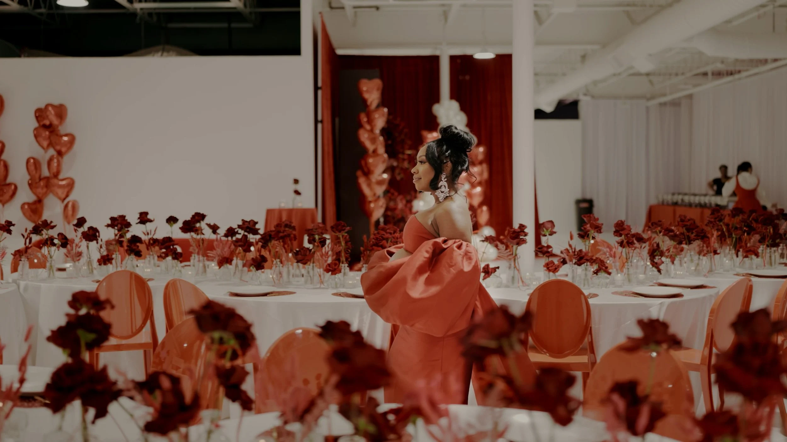 A woman in a red gown with off-the-shoulder sleeves stands among decorated tables at a celebration or event, surrounded by red and pink flowers and heart-shaped balloons.