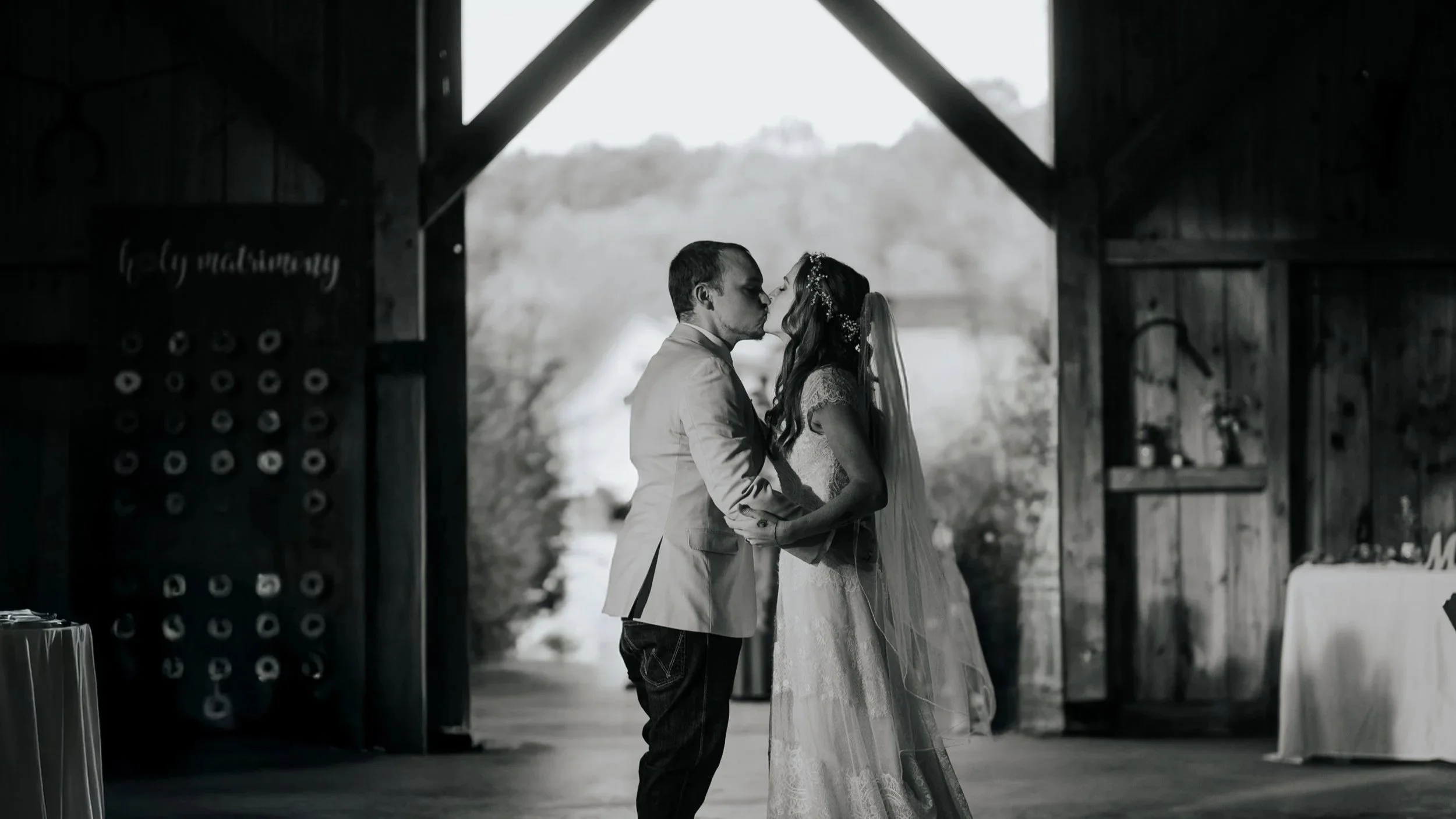A black and white photograph of a bride and groom sharing a kiss inside a rustic wooden barn, with trees and natural light in the background.
