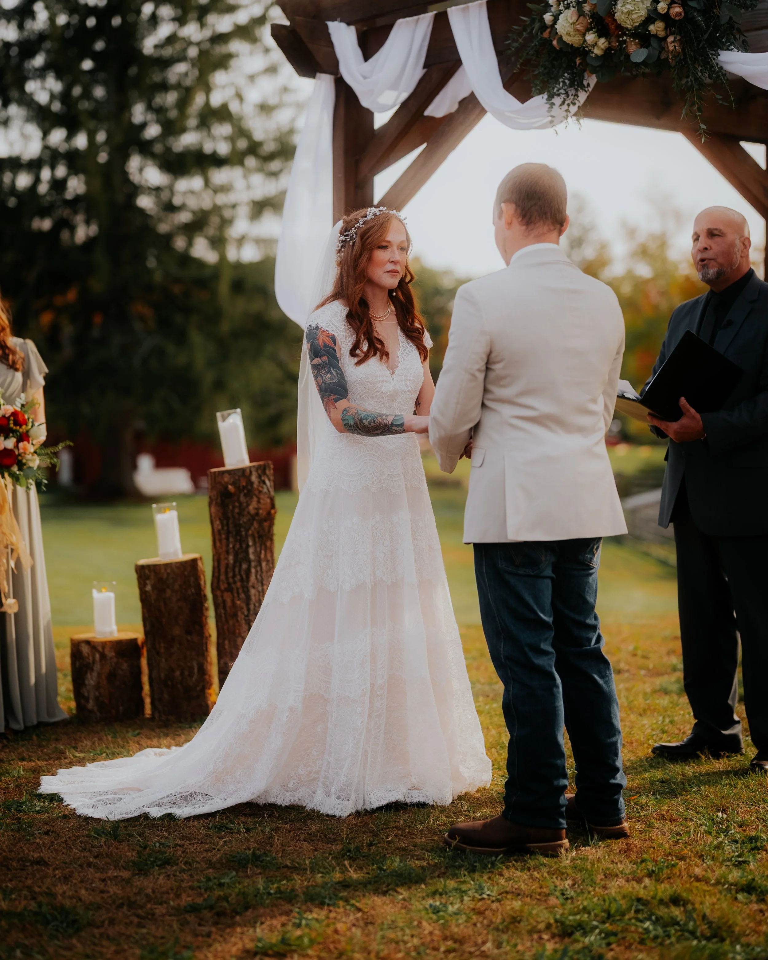 A couple getting married outdoors under a wooden arch decorated with white fabric and flowers, with an officiant present. The bride has red hair and tattoos, wearing a white lace gown, while the groom is in a white jacket and jeans.