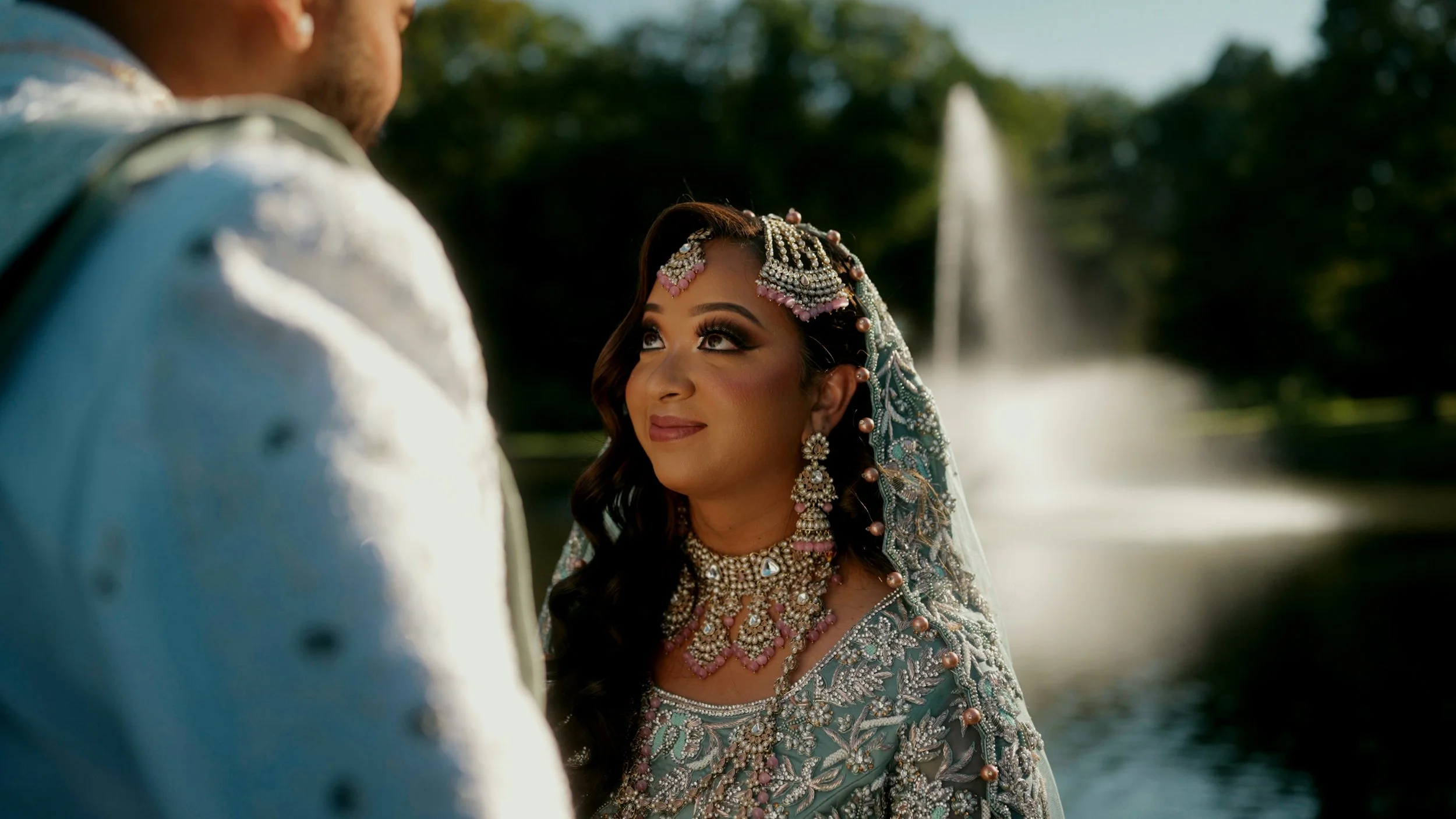 A woman dressed in traditional Indian wedding attire with jewelry, looking at a man, with a waterfall in the background.