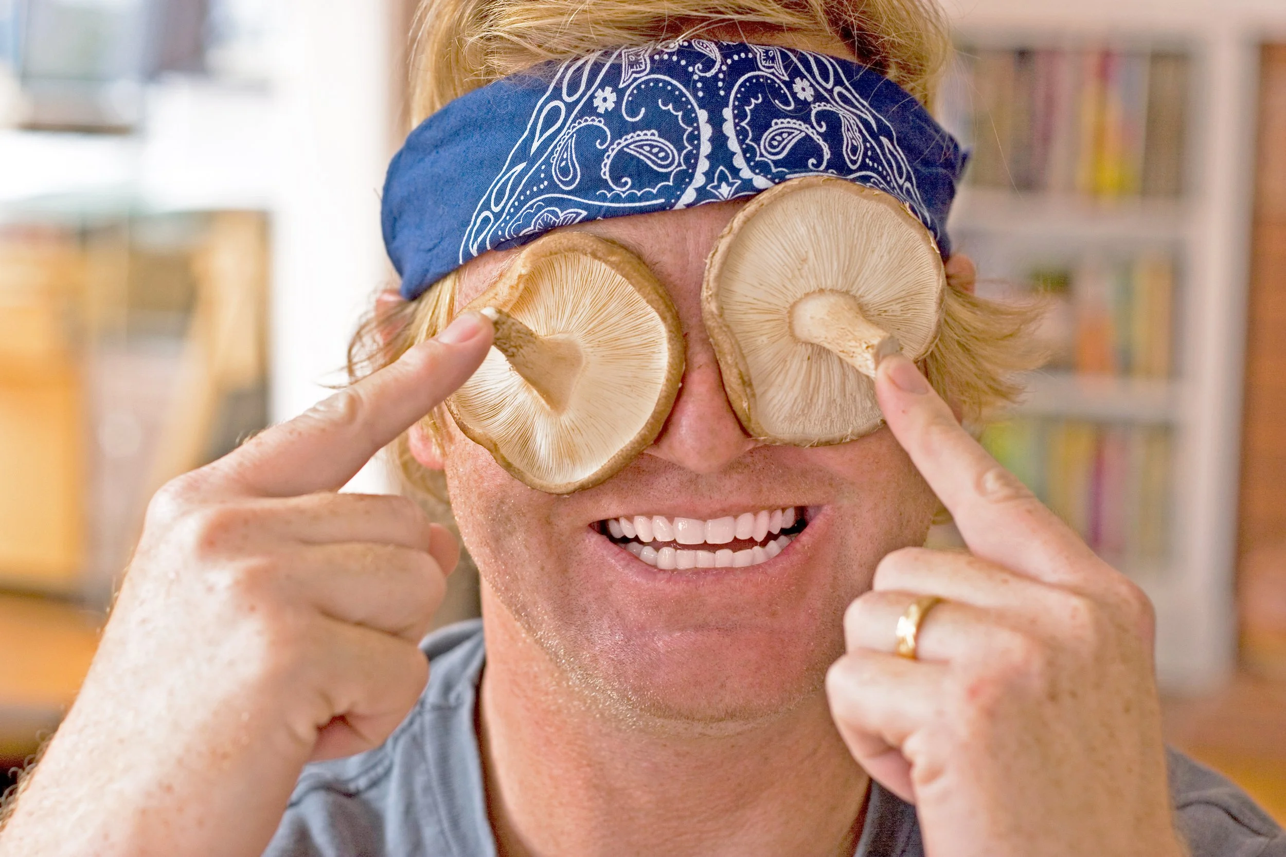A person with a blue bandana over their eyes, holding mushroom slices over their eyes, smiling with their teeth showing.