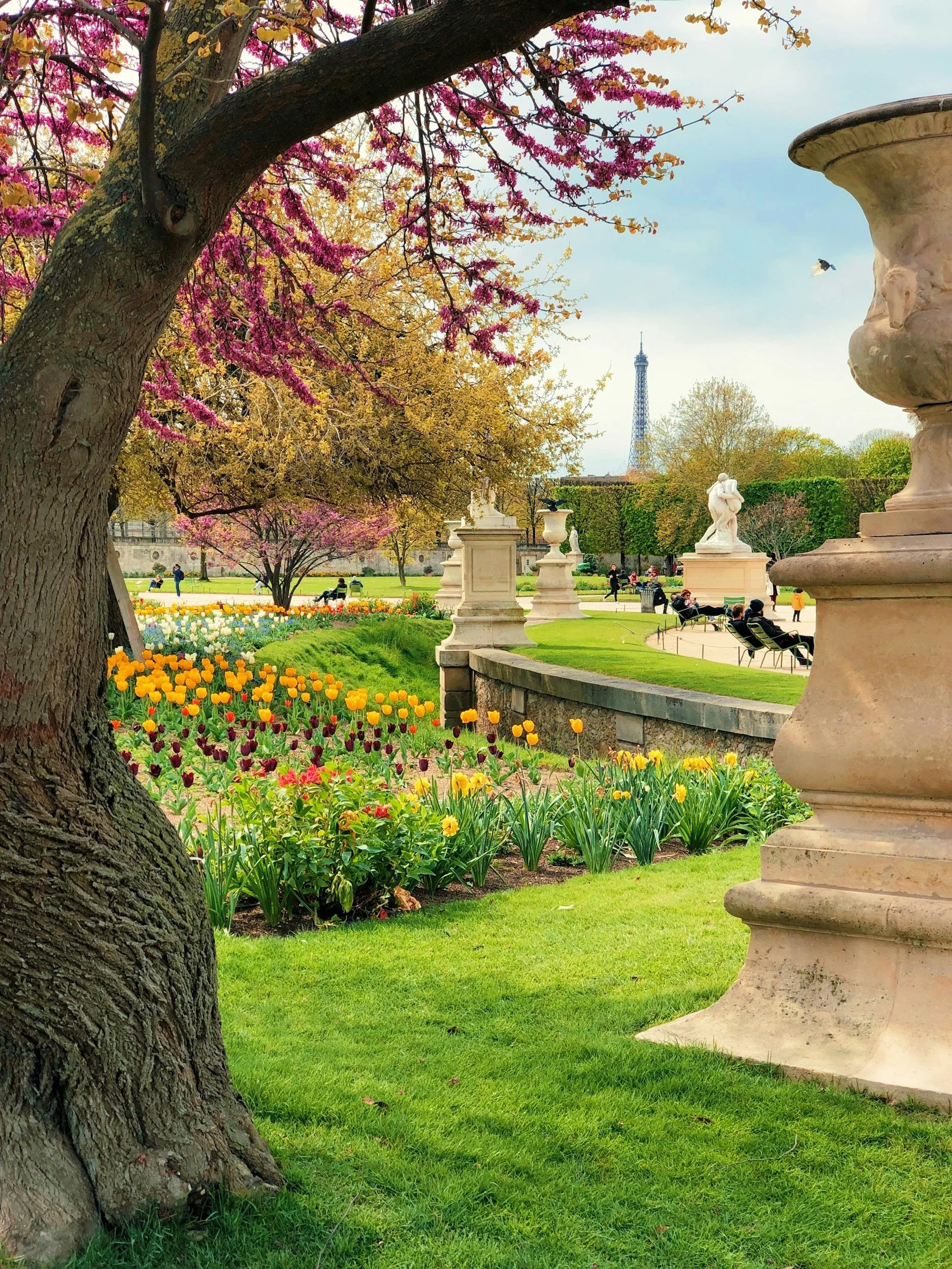A lush park scene with blooming pink and yellow trees, colorful tulips, green grass, and sculptures, with the Eiffel Tower visible in the background.