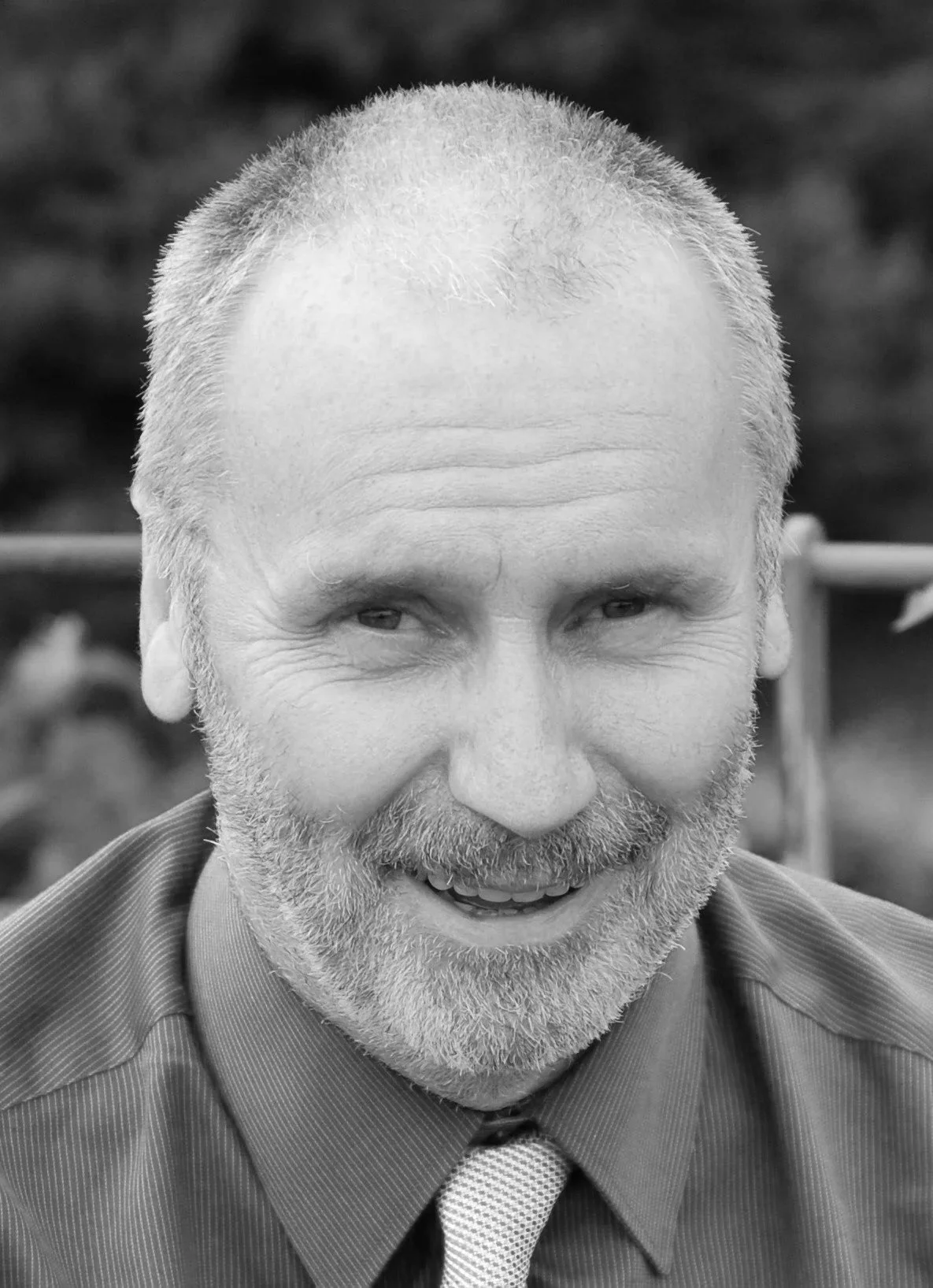 Close-up black-and-white photo of a smiling man with short hair, a beard, wearing a collared shirt and tie.