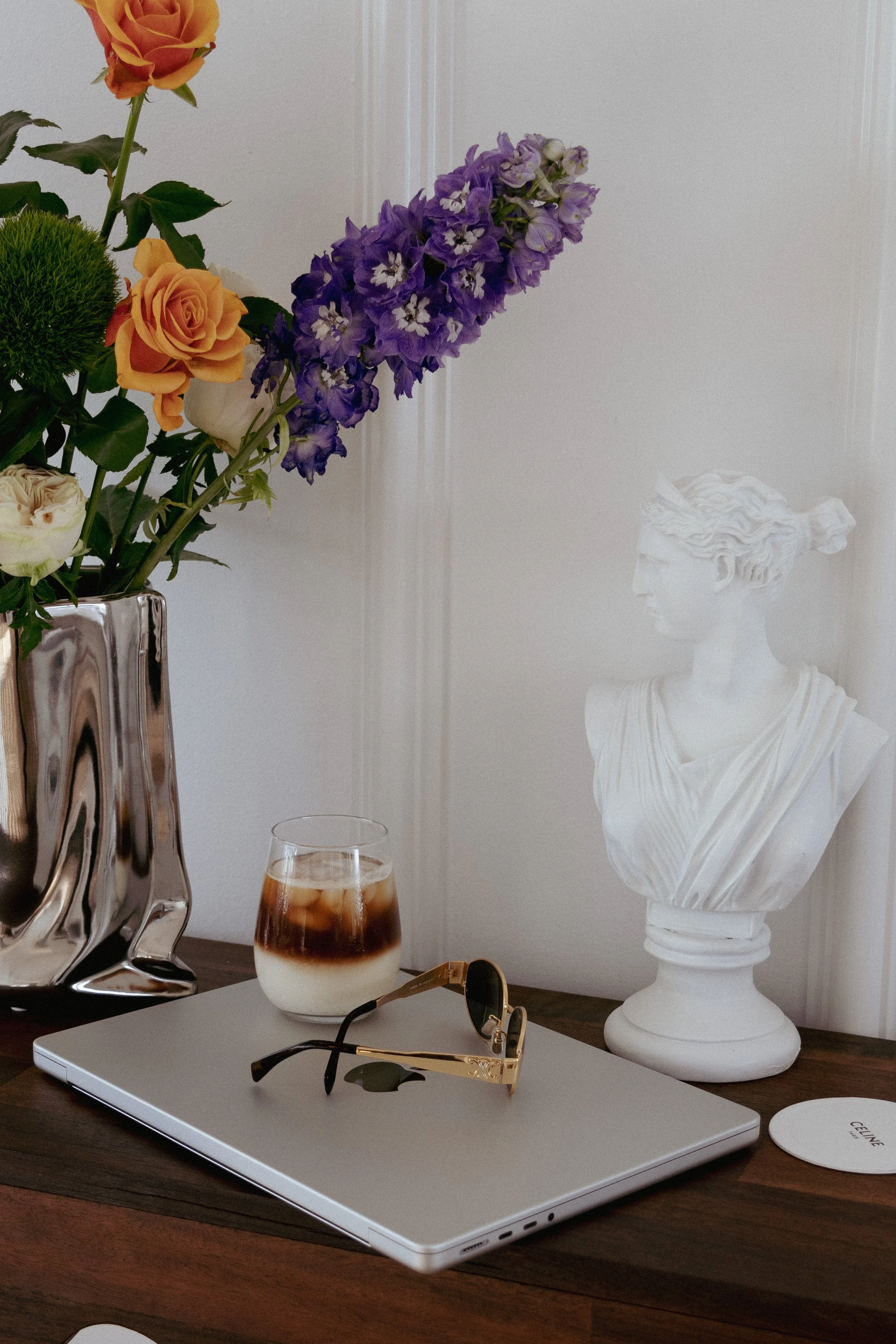 A wooden surface with a laptop, sunglasses, a glass of iced coffee, a floral arrangement with orange roses and purple flowers in a silver vase, a white marble bust sculpture, and a white circular object with text.