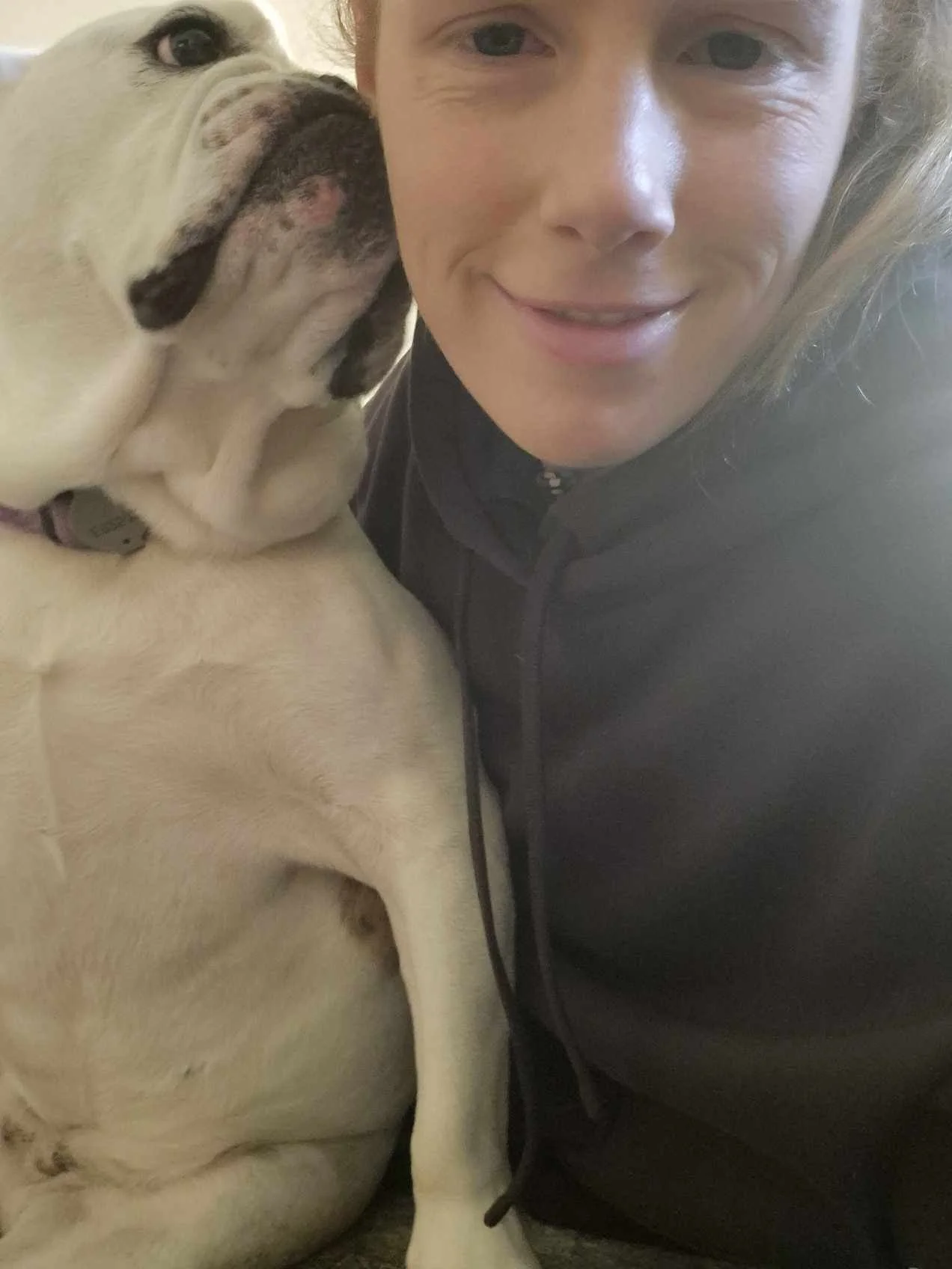A woman taking a close-up selfie with her cream-colored bulldog, who is licking her face.