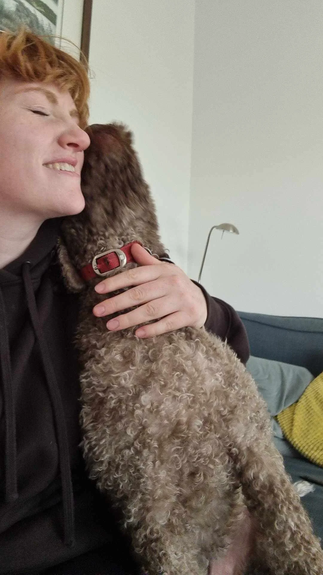 A woman with red hair smiling as she holds a curly-haired dog close to her face indoors.