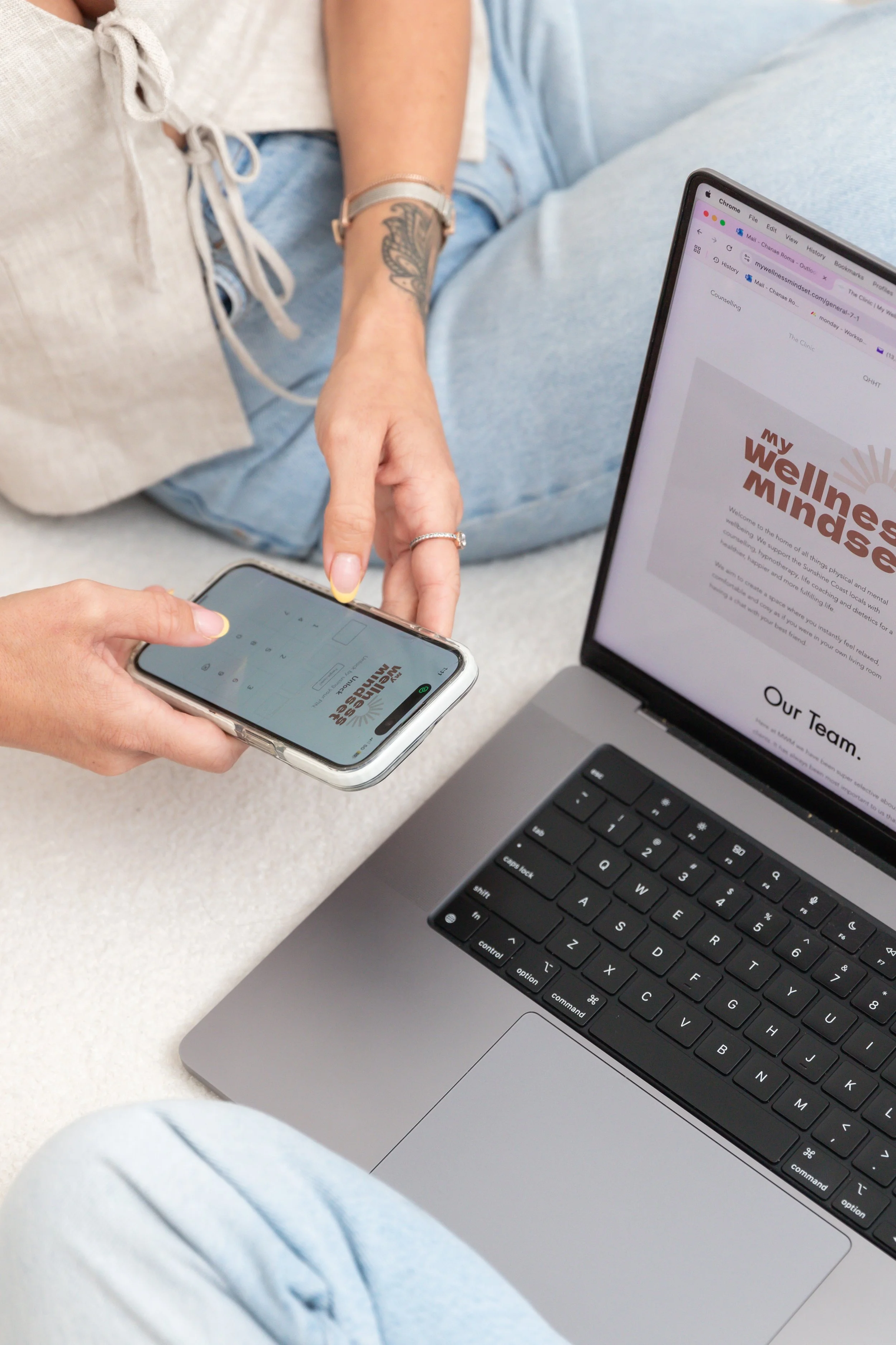 Person holding a smartphone displaying a website with the text 'My Wellness Mindset'. A laptop nearby shows the same website, with a visible section titled 'Our Team'.